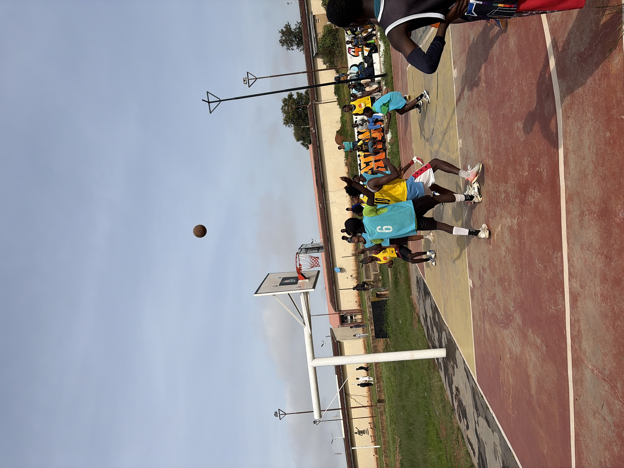 This image depicts an outdoor basketball game on a court in Jos, Nigeria. Several young men are actively playing, with some in mid-action, jumping or running, while others are observing from the sidelines. One individual is in the process of shooting a basketball, which is captured in mid-air. The court is marked with red and yellow surfaces, and a basketball hoop with a net is visible. In the background, there are buildings, trees, and overcast sky. A group of people are seated on a bench to the right of the frame. The scene suggests a casual game or practice session occurring in a public or community space.