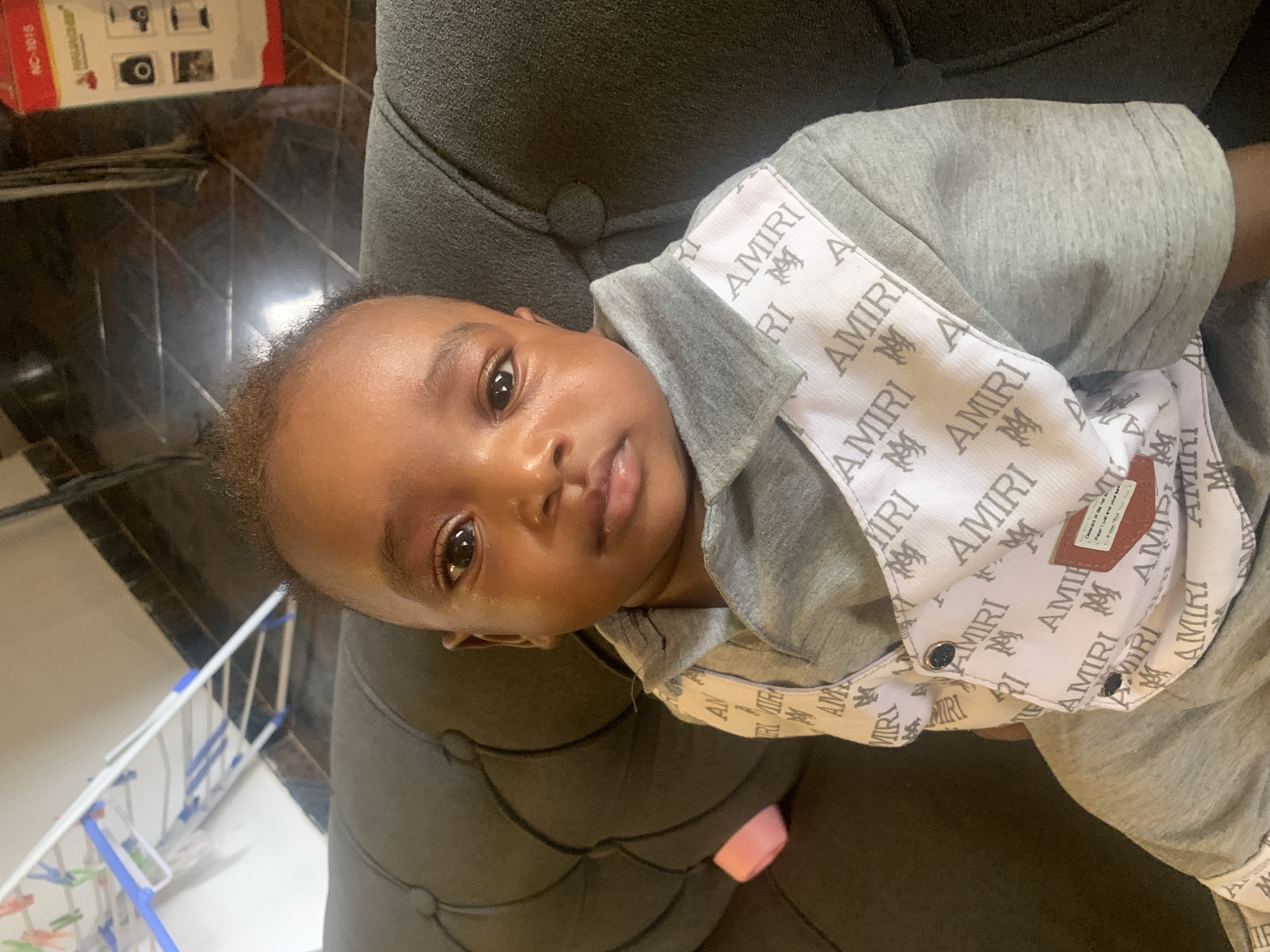 A close-up shot depicts a baby lying on a dark grey upholstered surface, possibly a couch or chair. The baby is wearing a grey outfit with the name "AMIRI" printed repeatedly on a white fabric lining or bib. The baby has dark skin, brown eyes, and short dark hair. The background is somewhat blurred, but a brown and black tiled floor is partially visible, along with a box labeled "NC-1015" and a drying rack with blue and white plastic elements. The scene is indoors. There is no specific indication of the location being Barakin Akawo, Nigeria.