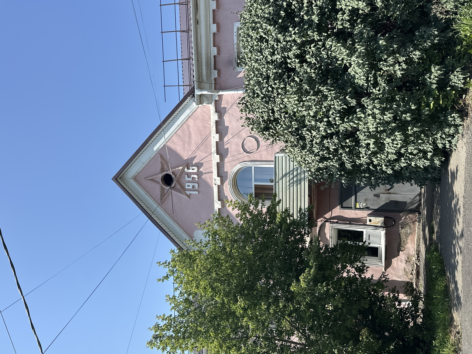 The image captures the facade of a pink building under a clear blue sky. The building features a triangular gable with the year "1956" prominently displayed in white numbers, along with a star emblem. A rounded window with white trim is visible above a section of green corrugated metal siding. To the left of the building, a large green tree stands, and to the right, a dense green bush partially obscures the lower part of the structure. In front of the building, a dark gray door is visible, next to a window with white trim. A conduit runs vertically along the wall near the window. The foreground shows a paved area, likely a sidewalk or road, with some grass growing along the edge. Overhead, several dark electrical wires crisscross the sky. The architectural style suggests a building from the mid-20th century, common in urban areas. This building is located in Biysk, Russia.