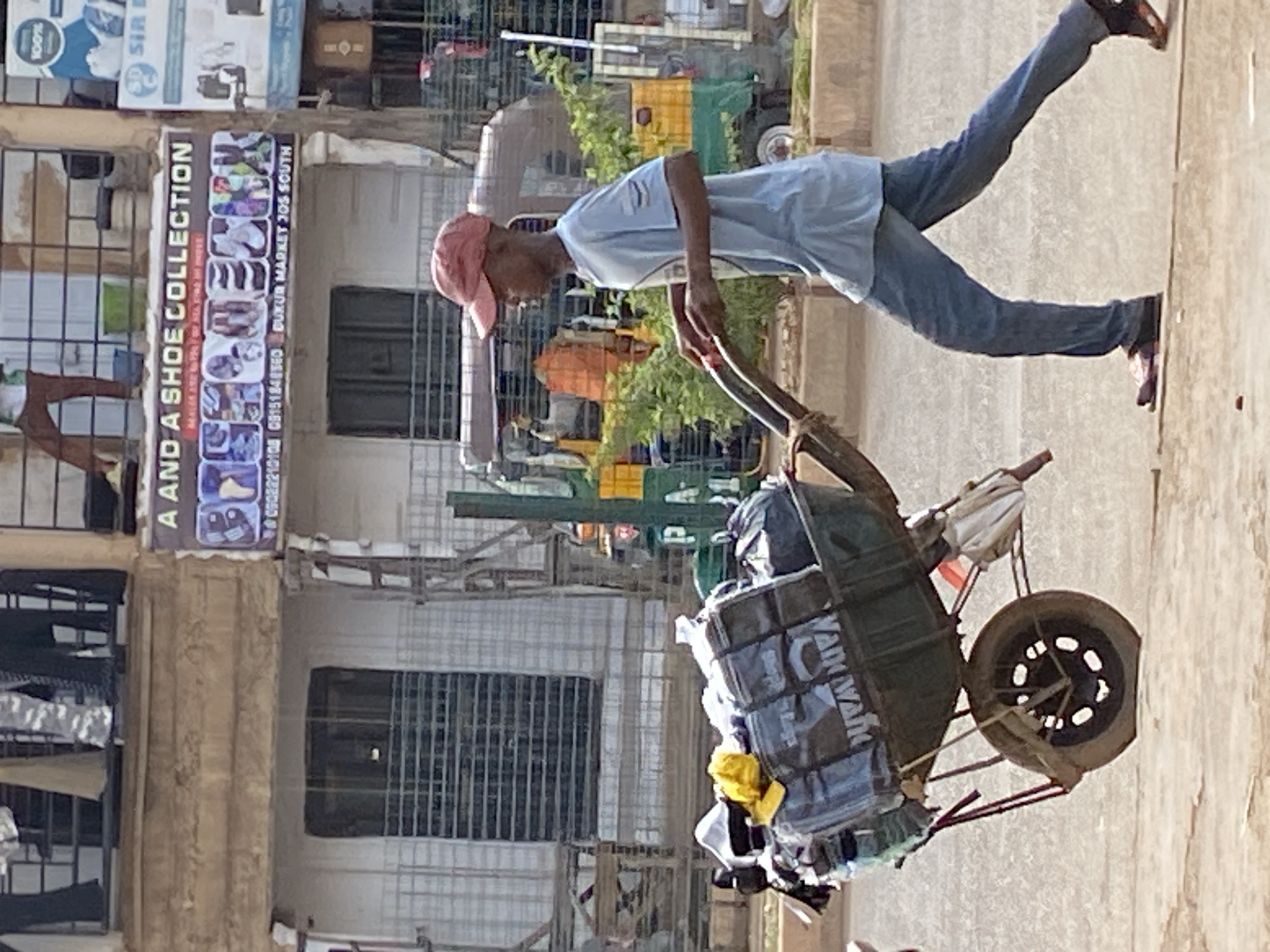 A young Black male wearing a light blue t-shirt, blue jeans, a red baseball cap, and brown and red sneakers is pushing a wheelbarrow loaded with bags and various items down a street in Bukuru, Nigeria. The wheelbarrow is dark gray with a single wheel in the front. The bags are wrapped in plastic and some have text. A yellow object is visible on top of the bags. Behind the man, a store with a banner that reads "A AND A SHOE COLLECTION" is visible. The store has a sign with images of shoes. The street appears to be paved and is lined with buildings. There are trees and other vehicles in the background.