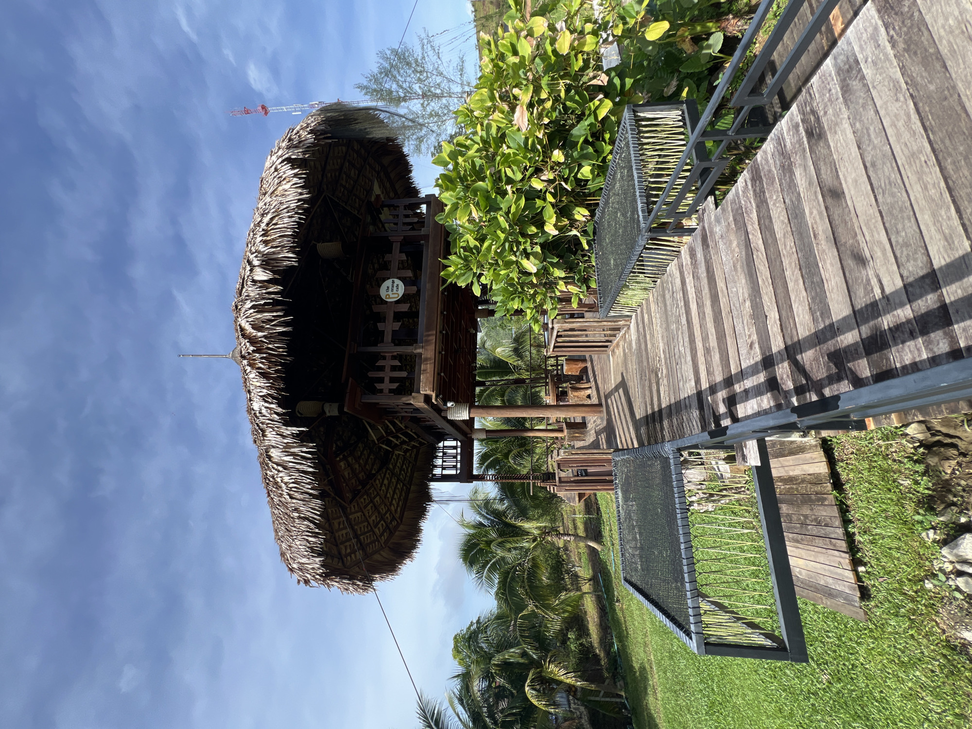 The image depicts an outdoor scene with a thatched-roof structure and a wooden walkway in Tambon Pa Kae Bo Hin, Thailand. The structure appears to be a pavilion or hut with intricate wooden carvings and a woven thatched roof. The walkway is made of weathered wooden planks and is lined with a modern metal railing that incorporates a mesh material. Lush green vegetation, including palm trees and leafy plants, surrounds the area, suggesting a tropical environment. The sky is a clear blue with some wispy clouds, indicating fair weather. In the distance, a red and white construction crane is visible against the sky. There is no activity taking place, and no people are visible in the image. The scene evokes a sense of tranquility and relaxation, characteristic of a resort or natural getaway. A small, circular sign with text in a circular frame is attached to the wooden structure, but the text is illegible. The lighting suggests it is daytime, possibly mid-morning or afternoon, given the shadows cast by the railing and the structure.