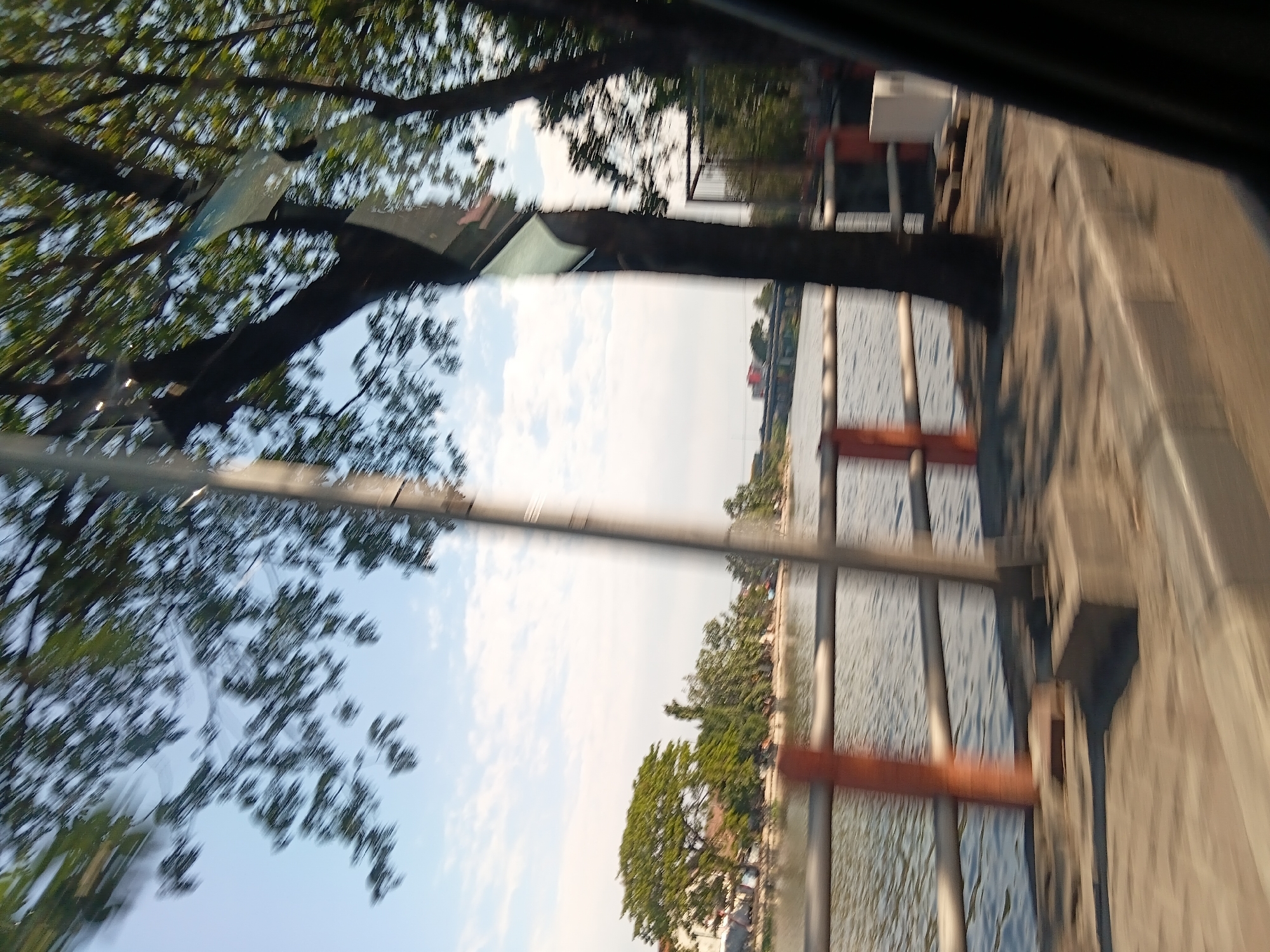 The image captures a blurry street-level view of a waterfront in Surabaya, Indonesia. The foreground features a blurred sidewalk with a curb, and a metal railing with orange vertical supports runs along the edge of the water. Beyond the railing, a body of water, likely a river or canal, is visible with ripples on its surface. Across the water, a tree-lined bank is seen with some buildings in the distance. A bridge can be observed spanning the water further down. Several trees are present in the scene, some in the foreground casting shadows, and others on the opposite bank. The sky is partly cloudy with a bright, clear blue expanse. The overall impression is one of a dynamic, perhaps moving, perspective of a cityscape with natural elements.