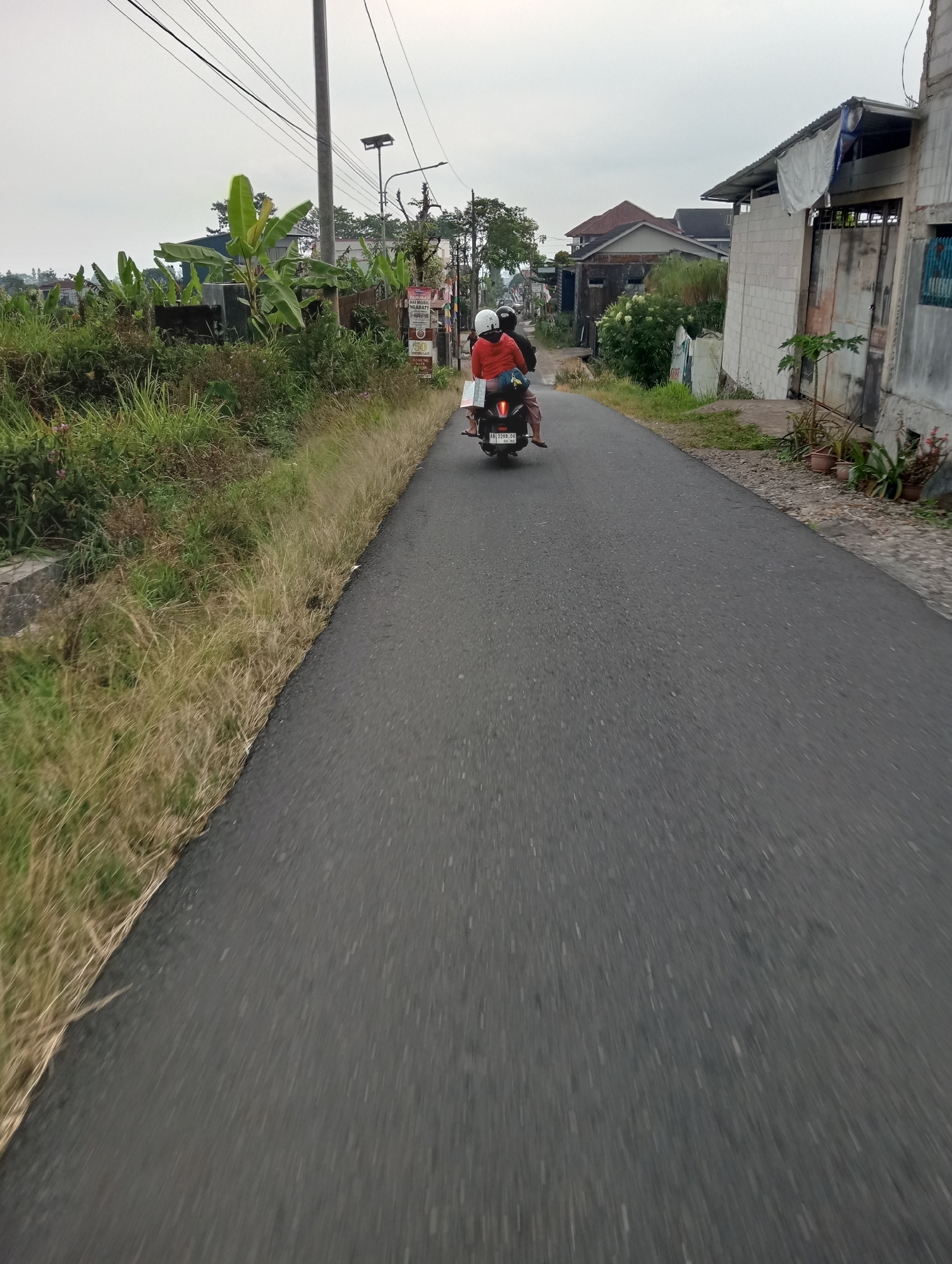 Two people, wearing helmets, ride on a dark-colored scooter down a paved road. The person in front is wearing a red jacket, and the person behind is wearing a pink shirt and patterned pants. The scooter has a license plate that reads "AB 3288 OV 03.20". The road is bordered by dry grass and some green vegetation on the left. On the right, there are buildings made of concrete blocks with corrugated metal roofs and some potted plants. In the background, there are more houses, trees, and a utility pole with electrical wires. The sky is overcast. The scene appears to be in a rural or suburban area of Pandansari, Indonesia.