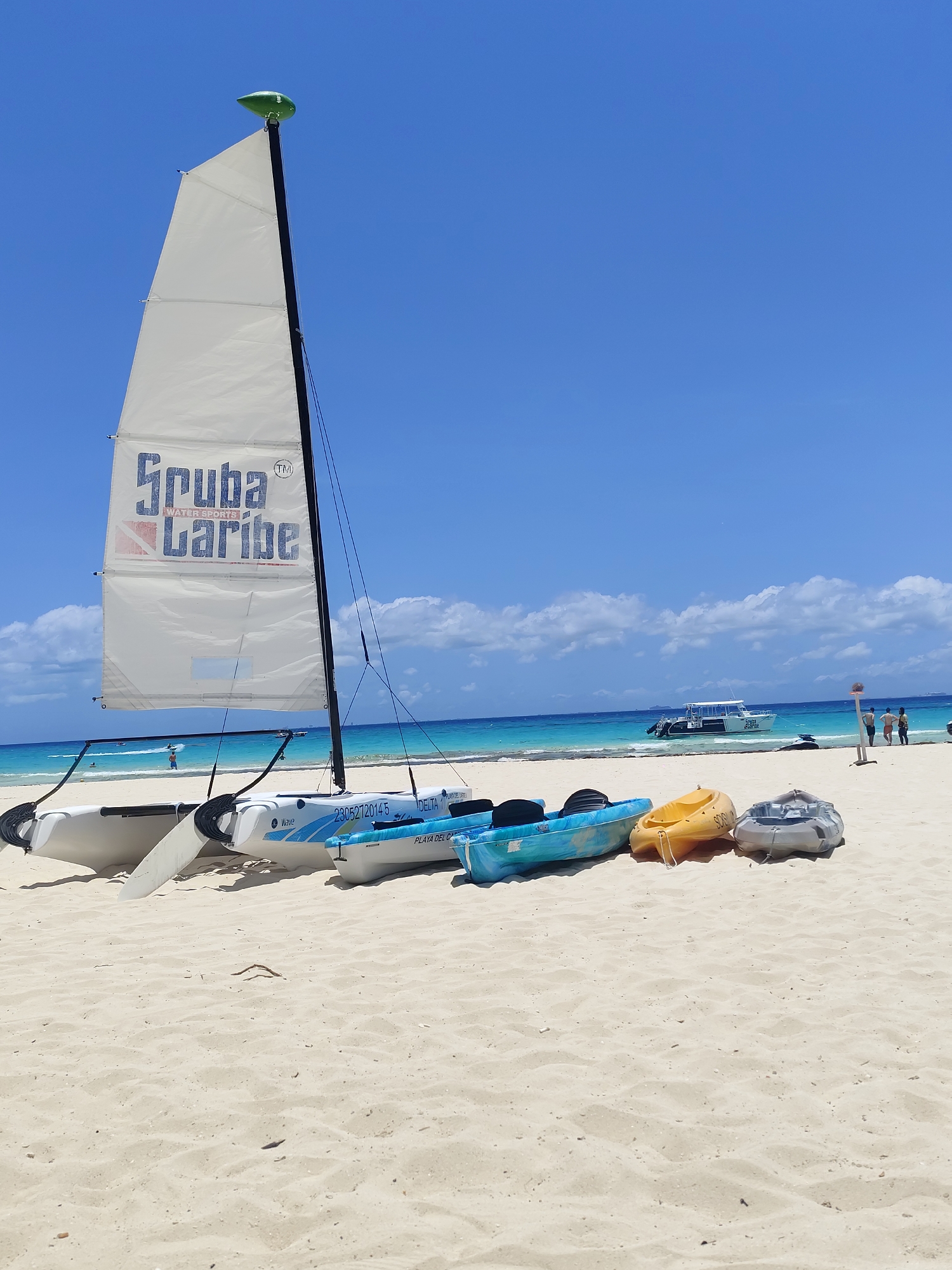 The image shows a bright, sunny day on a beach in Playa del Carmen, Mexico. In the foreground, several watercraft are beached on the white sand. A white catamaran with a tall sail bearing the words "Scuba Caribe" is prominent on the left. To the right are three kayaks of blue, yellow, and gray colors.
In the background, the turquoise water of the Caribbean Sea meets the horizon under a clear blue sky dotted with a few white clouds. A boat is visible in the distance on the water. On the right side of the background, a few people are standing on the beach. The overall scene depicts a calm day at a tourist beach, where water sports equipment is readily available.