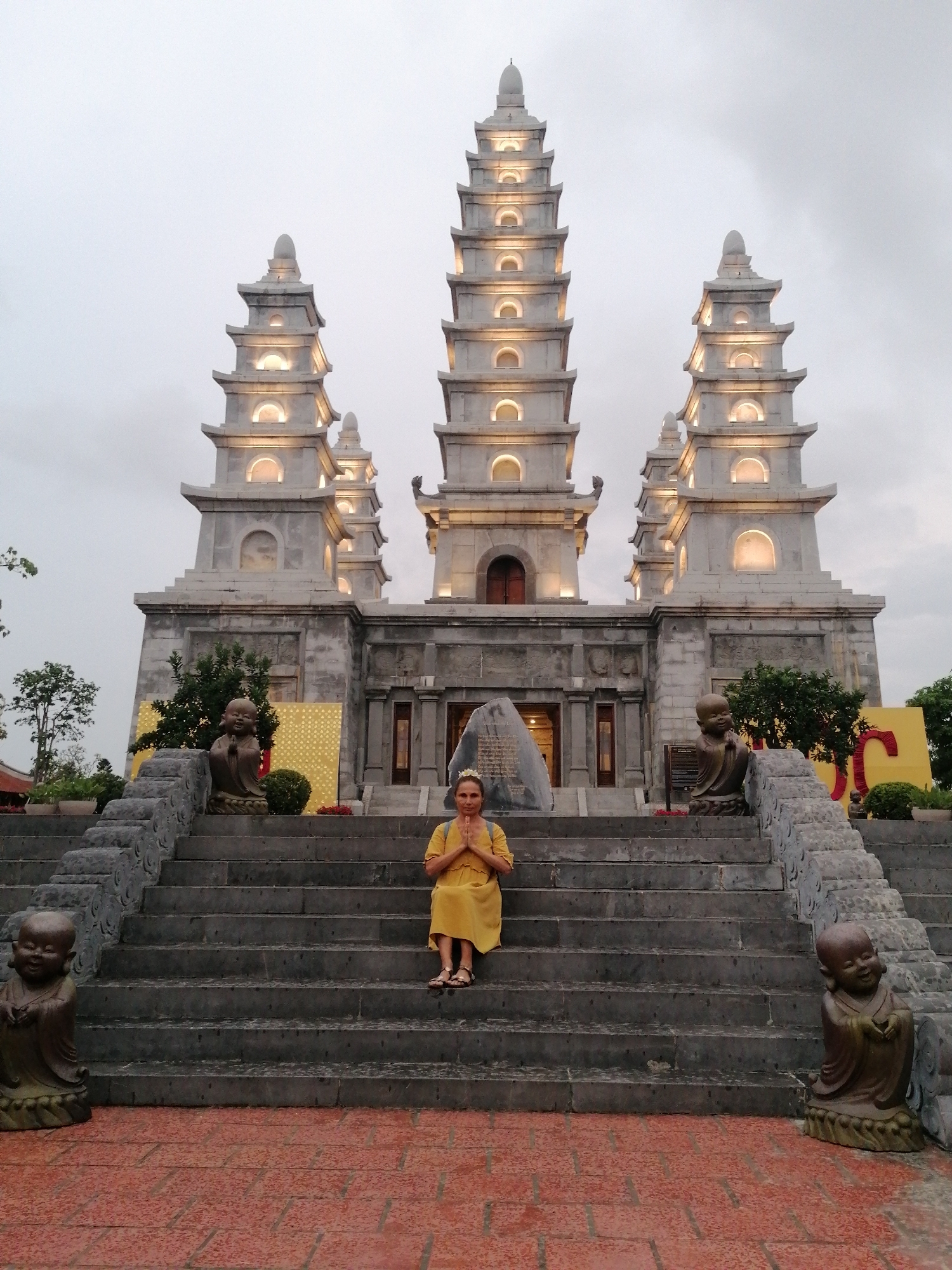A full shot shows a woman sitting on the stairs of a temple in the Hong Gai Ward of Vietnam. The woman is wearing a yellow dress, and has her hands clasped together in front of her. She is facing the camera. Behind her is a large stone temple with three tall towers. The towers are lit up with lights. The temple is on a hill, and there are stairs leading up to it. There are statues of children on either side of the stairs. The sky is cloudy. The foreground is a brick patio.
