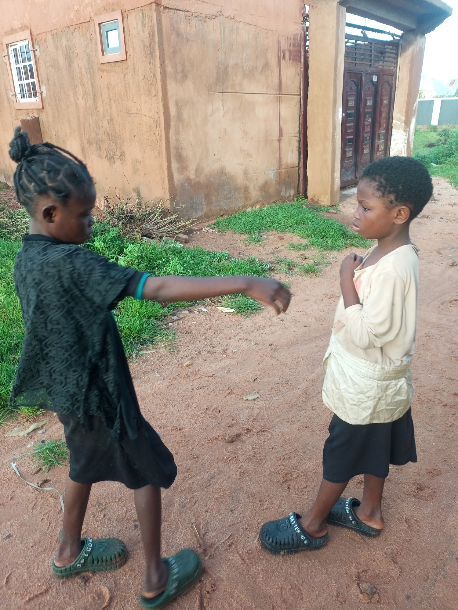 Two young children, a girl and a boy, are standing facing each other on a dirt path next to a light brown building in Tempure, Nigeria. The girl on the left, wearing a black lace dress and green clogs, has her left arm extended towards the boy. The boy on the right, dressed in a beige shirt and black shorts with black clogs, has his hands clasped in front of his chest. The building has two windows and a large wooden gate. Green grass and dry branches are scattered around the area.