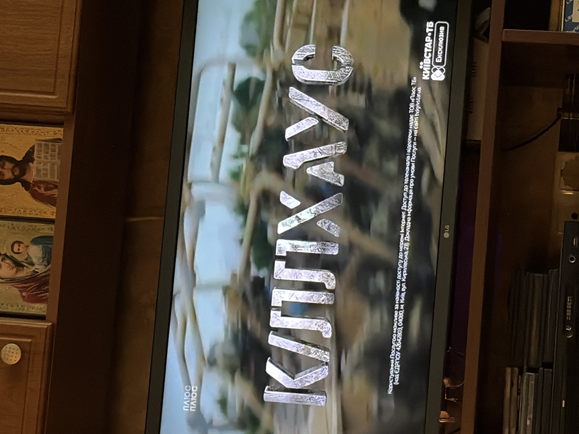 The image shows a close-up of a television screen displaying a program title in large, metallic, 3D Cyrillic letters. The letters appear to spell "КЦЛЛХАУС". Above the title, in the top left corner of the screen, is the word "ПЛЮС" repeated twice, in a smaller font. Along the bottom of the screen, there is text in Ukrainian, including a website address. In the bottom right corner of the screen, a logo for "КИЇВСТАР•ТБ" with the word "Ексклюзив" is visible. The background of the screen shows a blurred, outdoor scene, possibly a battlefield or construction site. Above the television, on a wooden shelf, are two religious icons. To the left of the icons is a small, round, decorative knob. The overall scene appears to be an indoor setting, likely a living room, with the television as the central subject.