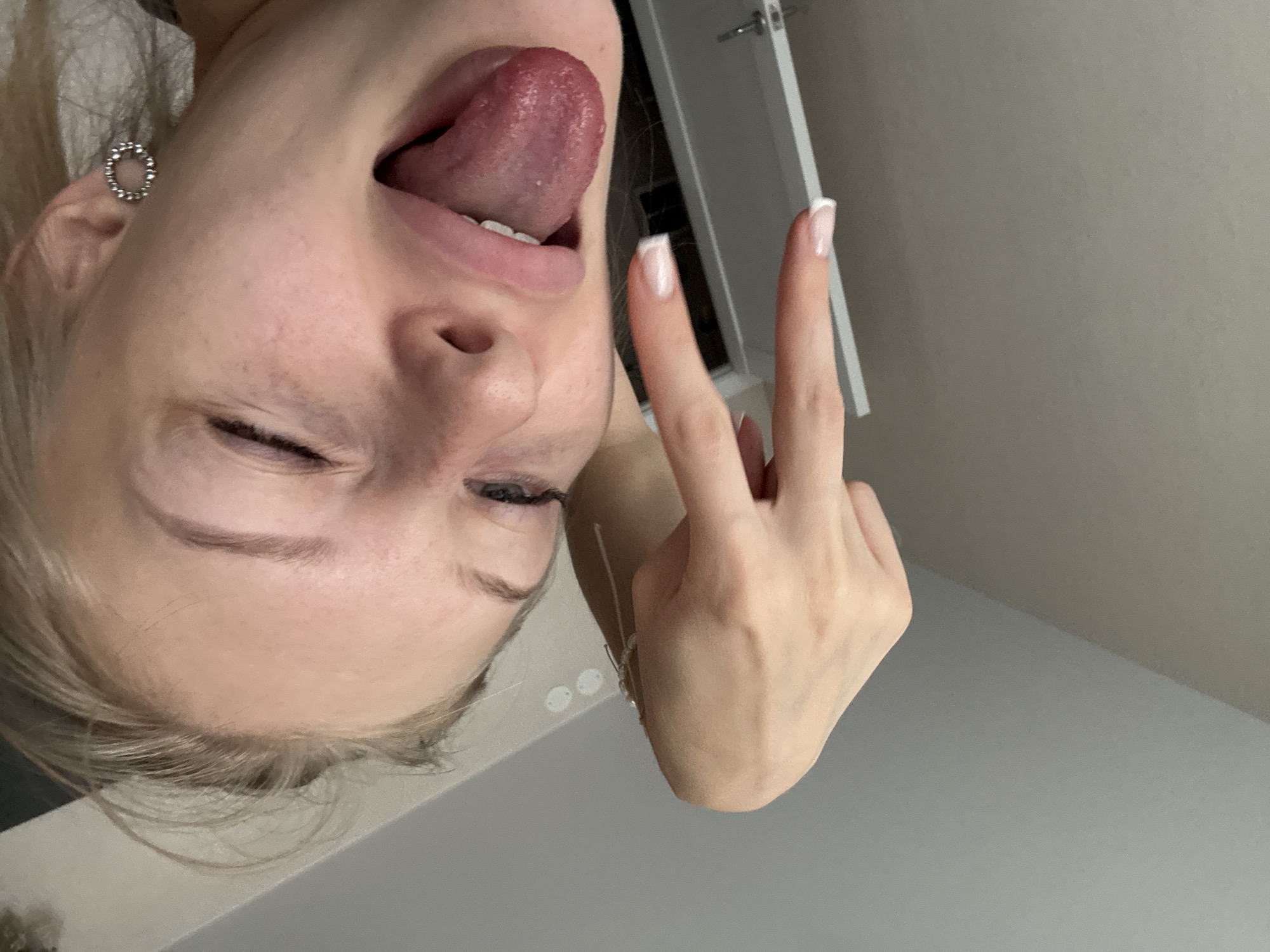 A young woman with blonde hair is shown in a close-up, selfie-style shot. She is winking and sticking her tongue out. Her left hand is raised, with her index and middle fingers extended in a "peace" or "rock on" gesture. She is wearing a silver earring in her right ear. The background appears to be an indoor setting with a wall and a corner of a ceiling visible. There are no identifiable landmarks that would suggest a specific location in Perm, Russia.