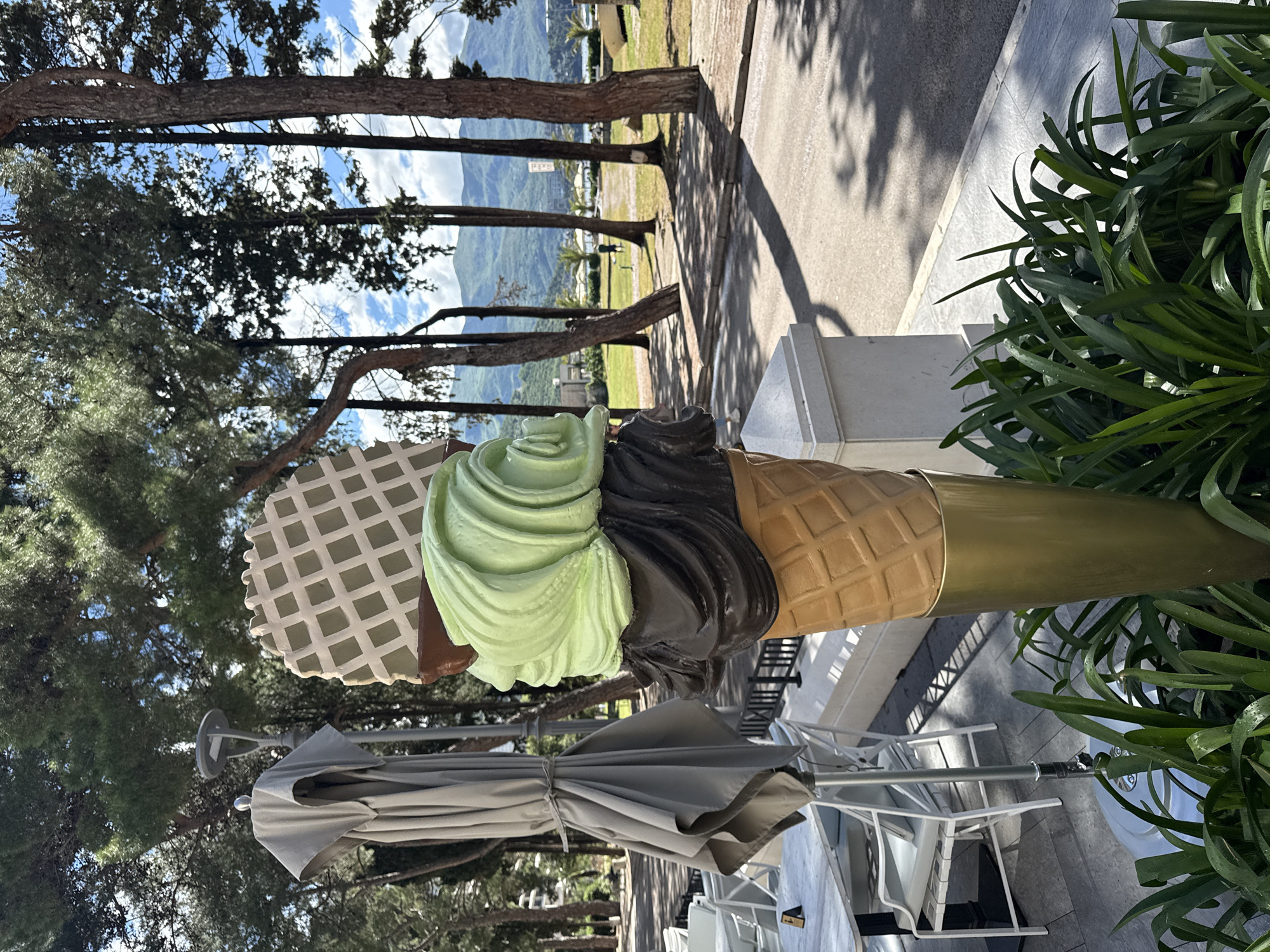 The image depicts a large, ornamental ice cream cone sculpture with three flavors: waffle cone, mint green, and chocolate. The cone is situated on a sidewalk in an outdoor cafe setting, indicated by the presence of white tables and chairs with a closed umbrella. In the background, there are trees and a view of mountains, suggesting the location is in a scenic area. Based on the architecture and landscape, this photo appears to be taken in Budva, Montenegro.