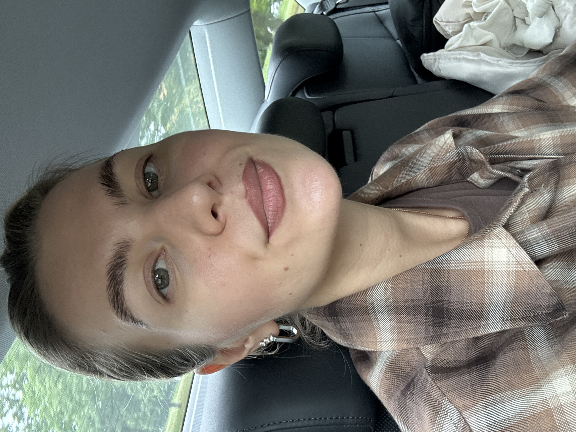 A woman is sitting in the back seat of a car. She is wearing a brown and white plaid shirt over a brown t-shirt and a thin rose gold necklace. She has on silver hoop earrings with three small stones. Her hair is pulled back. The car interior is visible behind her, with dark leather seats and a headrest. Outside the car window, trees and greenery are visible, suggesting an outdoor, possibly suburban or rural, environment. The overall lighting suggests it is daytime.