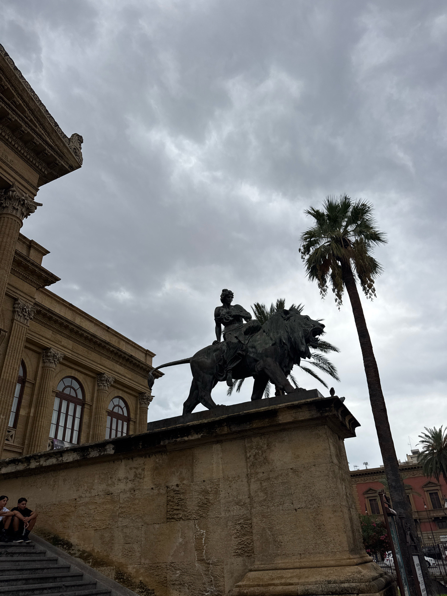 This image depicts a statue of a lion with a rider, positioned on a stone pedestal in front of a building with classical architecture. The building, likely the Teatro Massimo in Palermo, Italy, features large columns and arched windows. A tall palm tree is visible to the right of the statue, its fronds reaching towards a cloudy, overcast sky. Two young men are seated on a set of stairs in the lower left corner of the frame, facing the building. The scene appears to be an outdoor public space, likely a plaza or square.