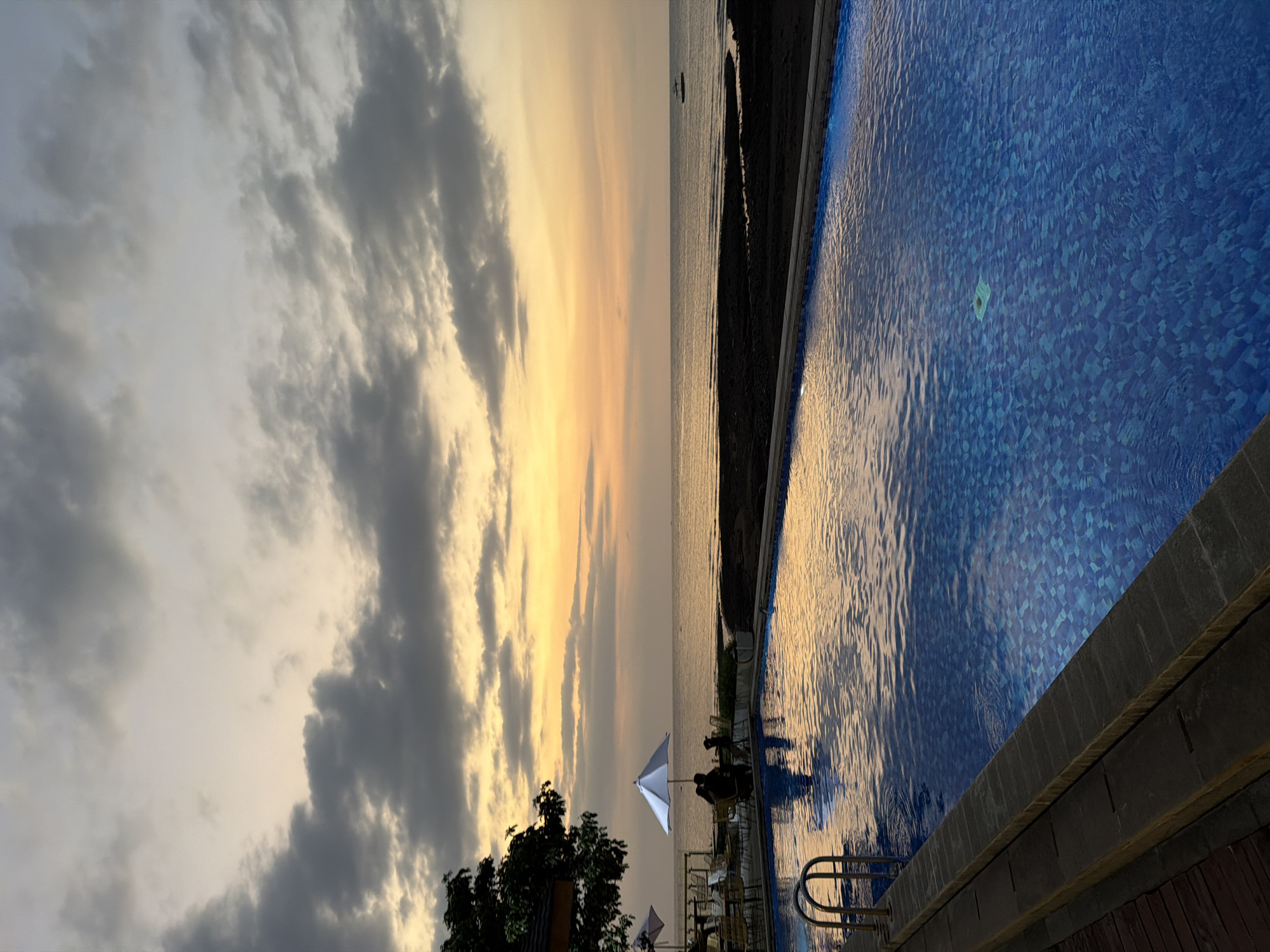 The image depicts a tranquil scene at sunset in Pasir Putih, Indonesia. A swimming pool with vibrant blue tiled depths occupies the foreground, reflecting the warm hues of the twilight sky. A metal ladder leads into the water from the left edge of the pool. Beyond the pool, a dark, sandy beach borders a calm sea, which stretches to the horizon. The sky above is a canvas of soft oranges, yellows, and grays, with scattered clouds creating a dramatic and serene atmosphere. In the distance, a small boat can be seen on the water. To the left of the pool, outdoor seating with chairs and tables is arranged, partially shaded by umbrellas. Silhouettes of a few individuals are visible, suggesting a relaxed outdoor dining or lounging experience. A lush green tree with dark foliage is situated in the upper left corner of the image, adding a touch of natural vibrancy. The overall impression is one of peaceful seaside relaxation at the end of the day.