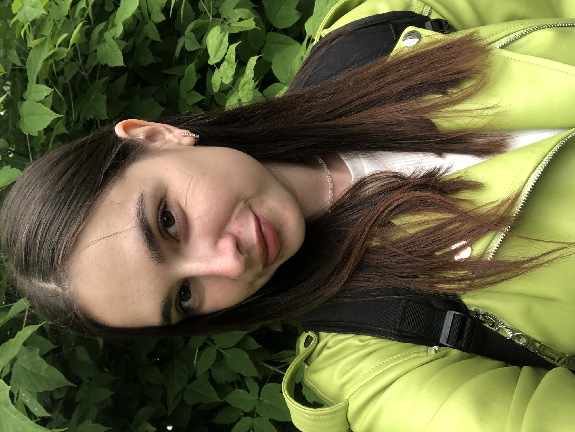 This is a portrait-oriented, slightly tilted, eye-level, medium shot of a young person in Chaikovsky, Russia. The subject has long, dark brown hair and is wearing a bright lime green jacket over a white top. A black strap with a buckle is visible on the jacket, likely part of a backpack or bag. A delicate, sparkling necklace is also visible around the subject's neck. The subject is looking towards the camera with a slight smile and their head tilted to their left. Their left ear is visible, adorned with a small, sparkling earring. The background consists of lush, green foliage, suggesting an outdoor setting, possibly a park or garden. The lighting appears to be natural daylight, and there are no obvious weather cues. The overall mood is casual and relaxed. No visible text is present in the image.