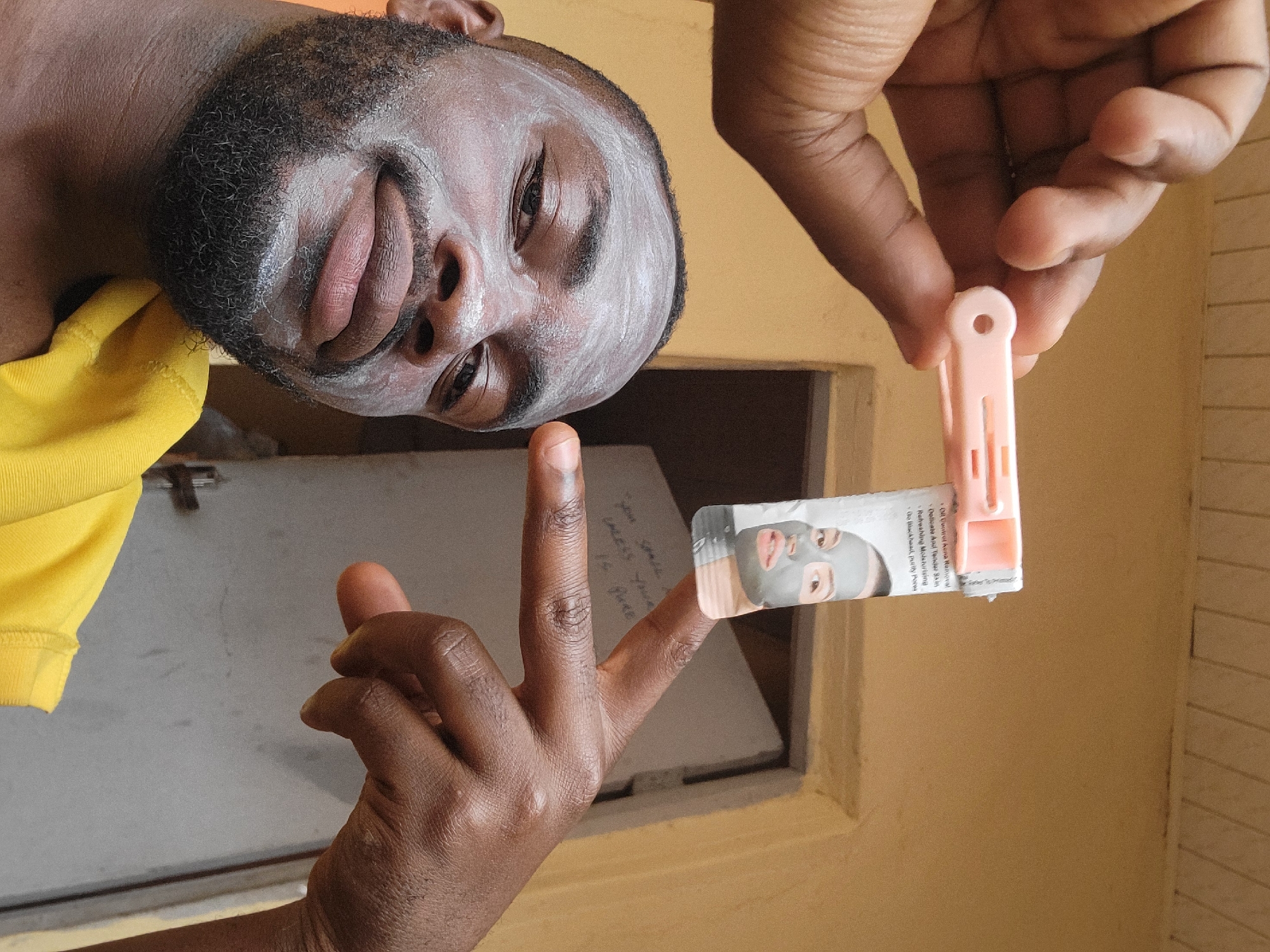 A medium shot captures a man with a white facial mask, contrasting his dark skin tone and beard, positioned in the upper-left of the frame. He is wearing a yellow shirt. His head is tilted slightly.
Below his face and positioned centrally is a partially visible hand with the middle and index fingers extended. Between these two fingers, a small piece of paper is held in place by a pink plastic clothespin. The paper has an image of a face wearing a similar facial mask. Text is visible on the paper, apparently instructions.
Above the extended fingers and towards the top right, another hand is visible.
A white surface with some writing can be seen behind the subject's middle finger. The background consists of a yellow wall.
There is no information to suggest that the image was taken in Jos, Nigeria.