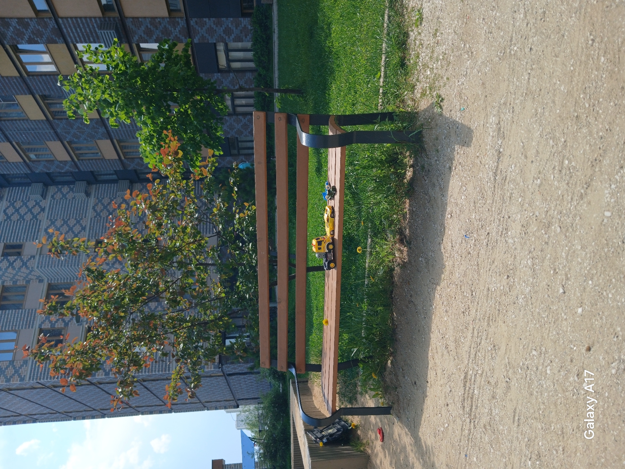 The image shows a park bench in Yekaterinburg, Russia. The bench is made of wood and metal, and is placed on a grassy area next to a gravel path. There are toy cars on the bench. Behind the bench is a building with multiple windows. A tree is visible in front of the building. The sky is visible at the bottom of the image, indicating a sunny day.