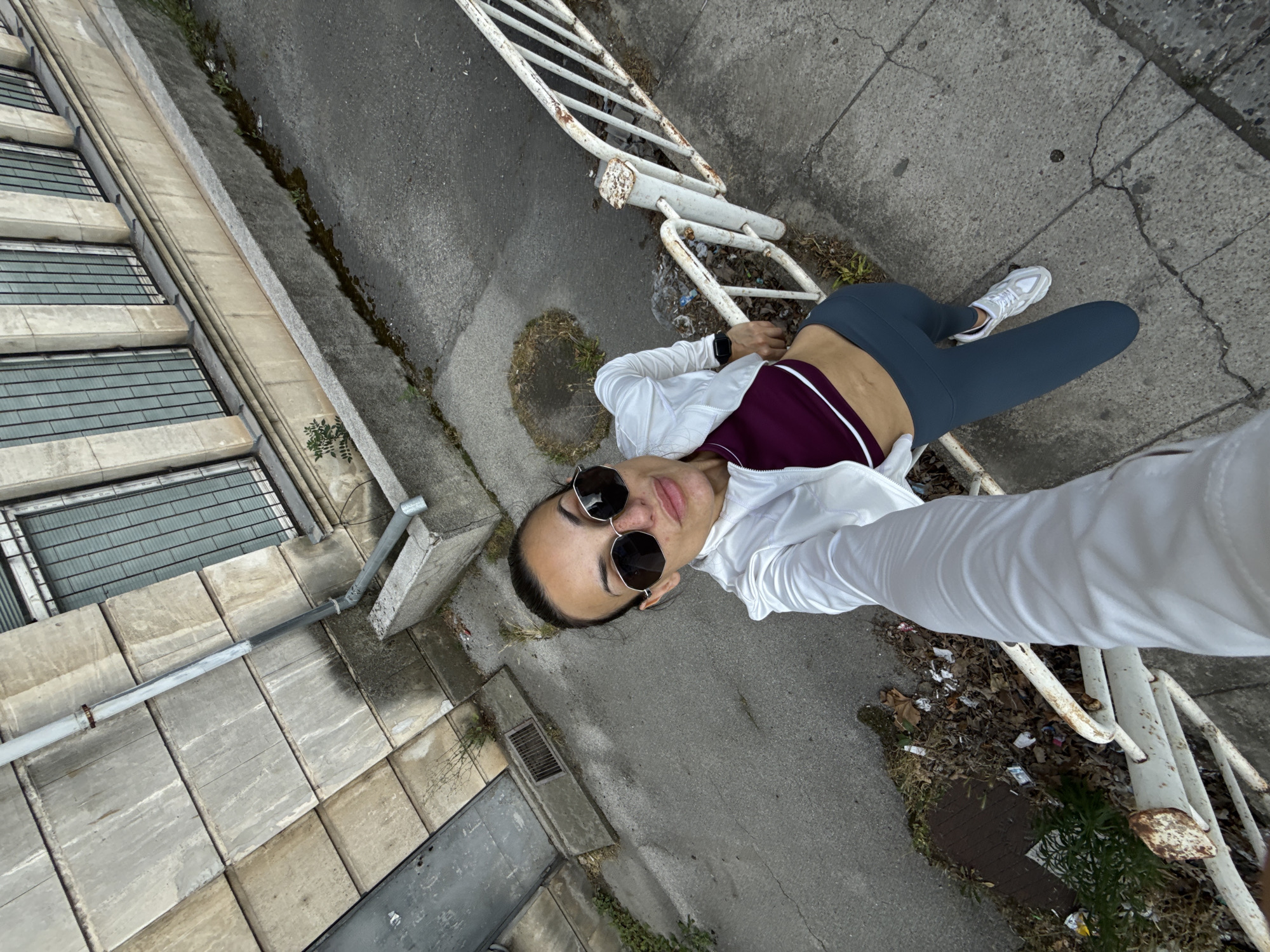 An overhead, high-angle selfie shows a woman in athletic wear standing on a sidewalk next to a rusty white metal railing. The woman, wearing sunglasses, a white jacket, a burgundy sports bra, and grey leggings, is holding her phone out to take the picture. She appears to be in an urban environment, with a concrete building and a street visible in the background. The building features large windows with metal bars and a grey downspout. The street is asphalt and shows signs of wear, with cracks and debris. The setting is consistent with the city of Pleven, Bulgaria, possibly a less maintained area.