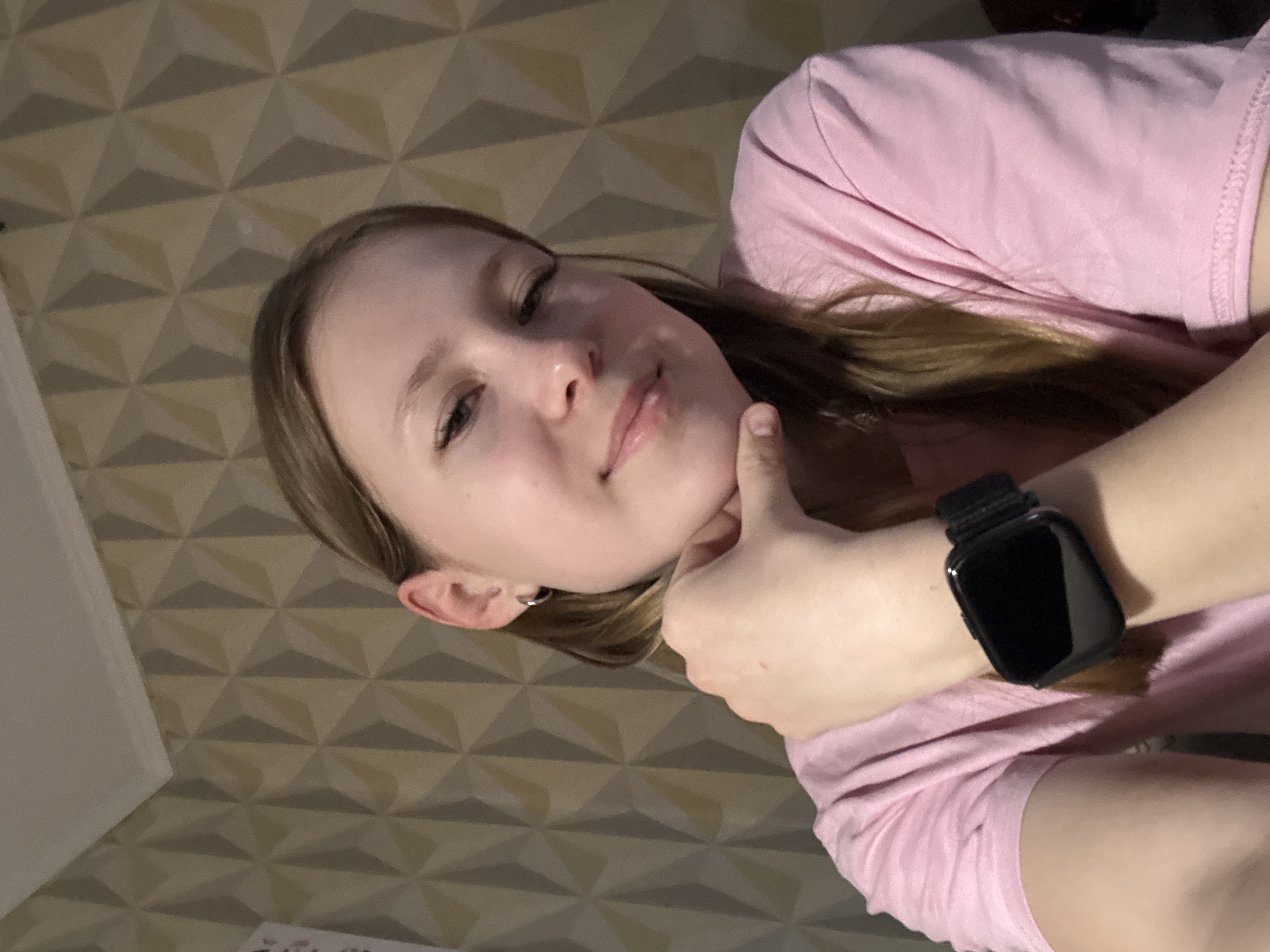 A young, light-skinned person with long, light-brown hair and pink clothing is looking directly at the camera. They have an earring in their left ear and are resting their chin on their right thumb. A black smartwatch is on their left wrist. The background is a patterned wall of three-dimensional triangles in neutral tones. The photograph appears to have been taken indoors. There is no verifiable evidence within this image to determine a specific location in Chaikovsky, Russia.