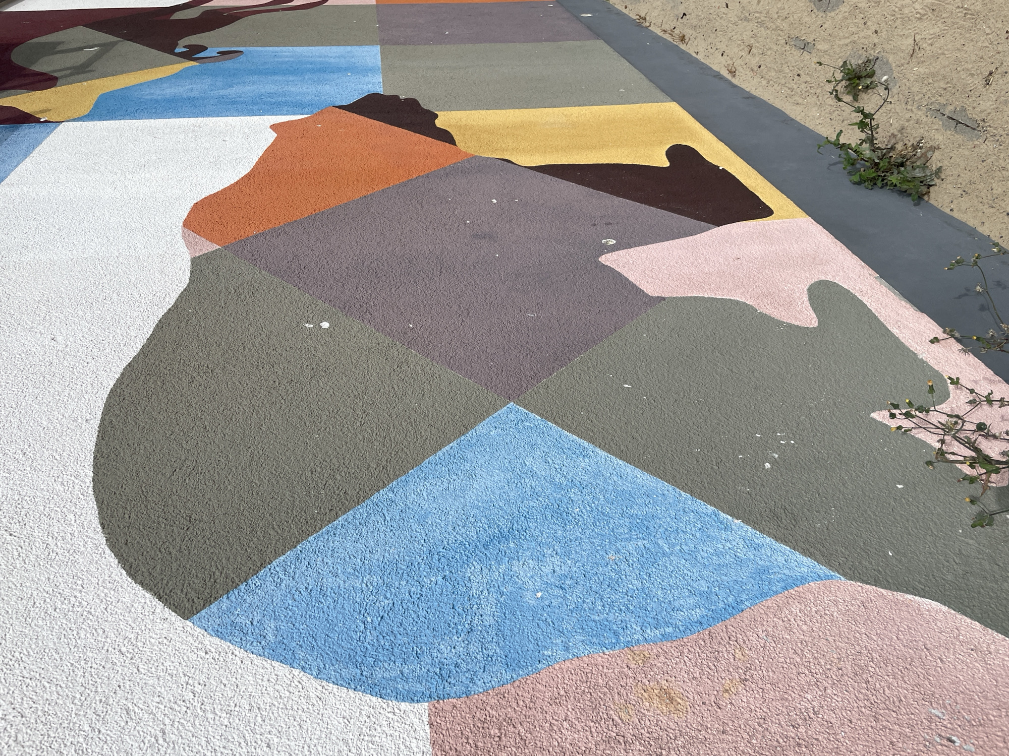 The image depicts a section of a colorful mural on an exterior wall in Bremerhaven, Germany. The mural features abstract geometric shapes and stylized human profiles in various muted colors, including white, light pink, light blue, gray, dark purple, orange, yellow, and dark brown. The wall has a textured, stucco-like finish. The lower part of the image shows a gray base along the bottom of the wall, with a concrete or paved surface beside it. Small green plants are growing out of cracks in the concrete near the wall. The scene is illuminated by natural daylight, casting shadows on the ground.