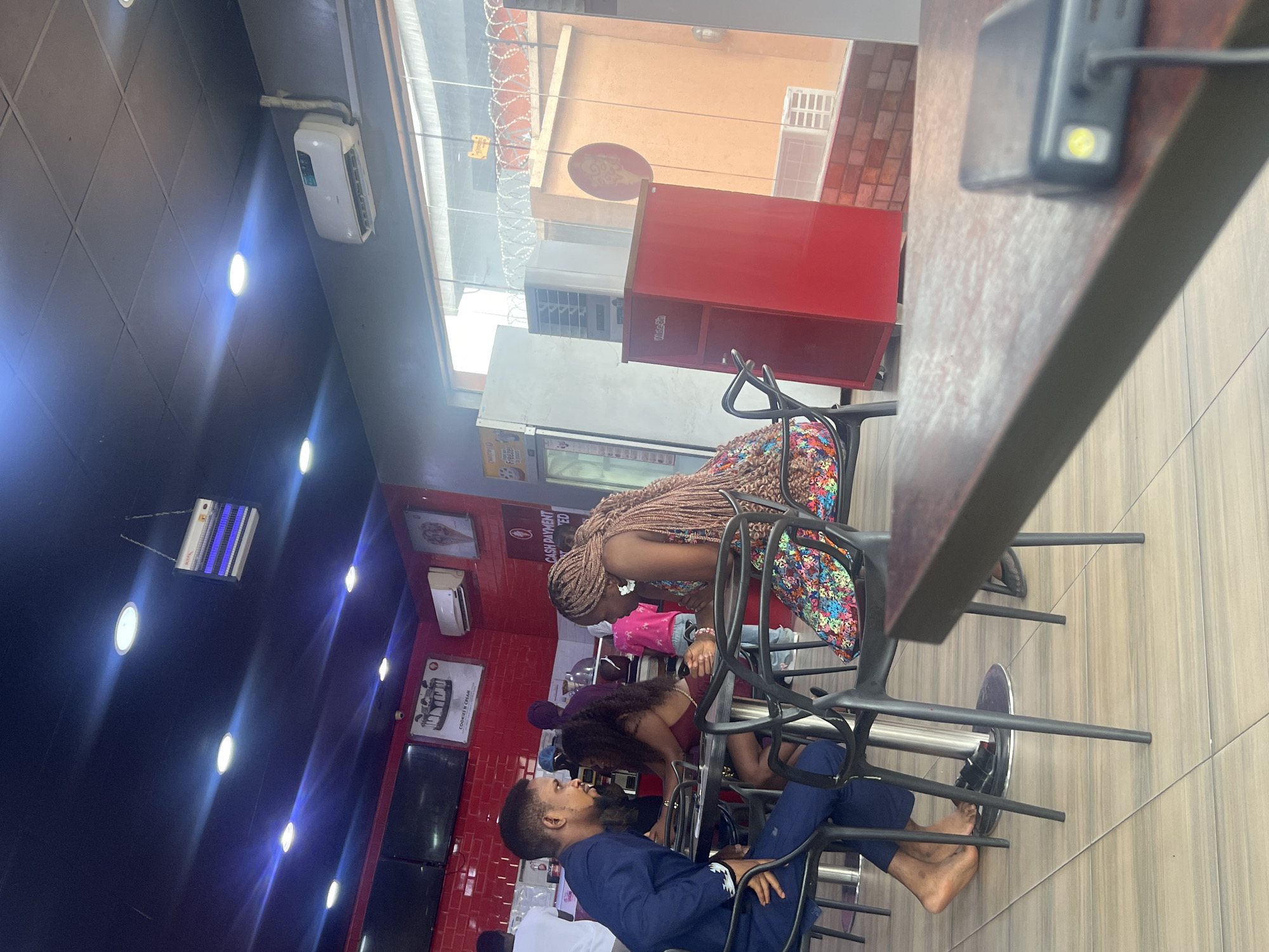 A group of people are seated indoors at a fast-food restaurant in Lagos, Nigeria. A woman with braids is sitting and leaning over a table. Another woman with dark hair is bent over near her. A man in a blue jacket and pants is also seated at the table. Other people are visible but less clearly. The restaurant has red walls, and there are advertising signs on the wall. The floor is tiled, and there is a table and chair. There are lights on the ceiling.