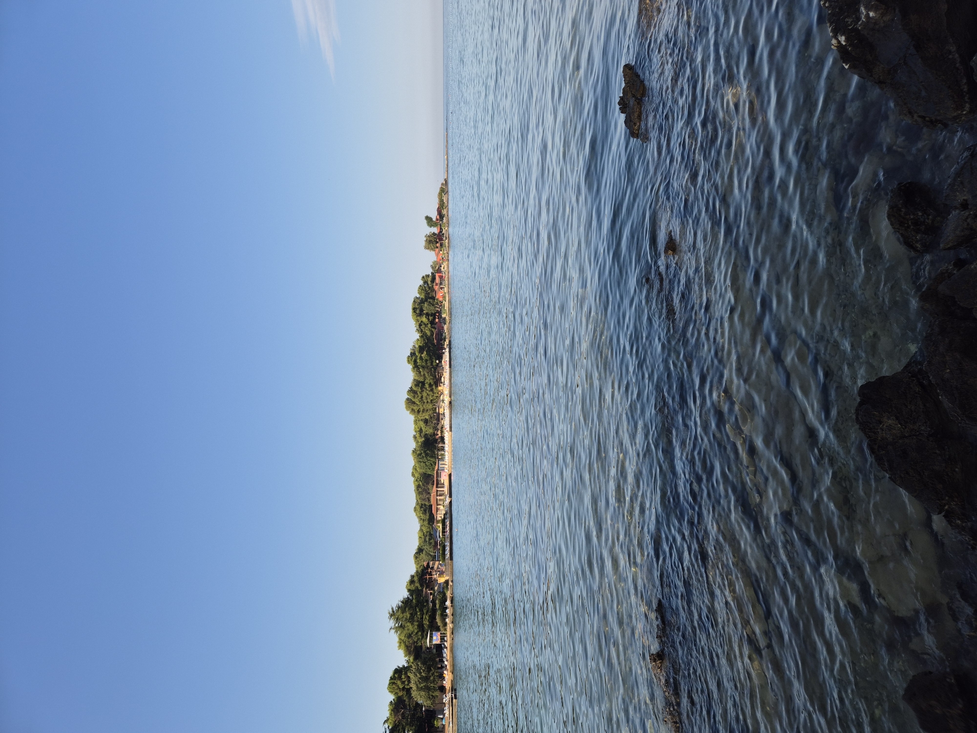 The image shows a serene view of the Adriatic Sea from the shore in Monterol, Croatia. The foreground is dominated by dark, rugged rocks partially submerged in the clear, rippling water. The water itself is a deep blue, transitioning to lighter shades closer to the shore, revealing the rocky seabed beneath.
In the mid-ground, a line of trees forms a verdant backdrop to the scene. Behind the trees, a cluster of red-roofed buildings and beach structures are visible, suggesting a resort or tourist area. A few scattered lounge chairs are visible on the beach, hinting at a leisurely atmosphere. The sky above is a clear, bright blue, devoid of clouds, indicative of a sunny day. The overall impression is one of tranquility and natural beauty.