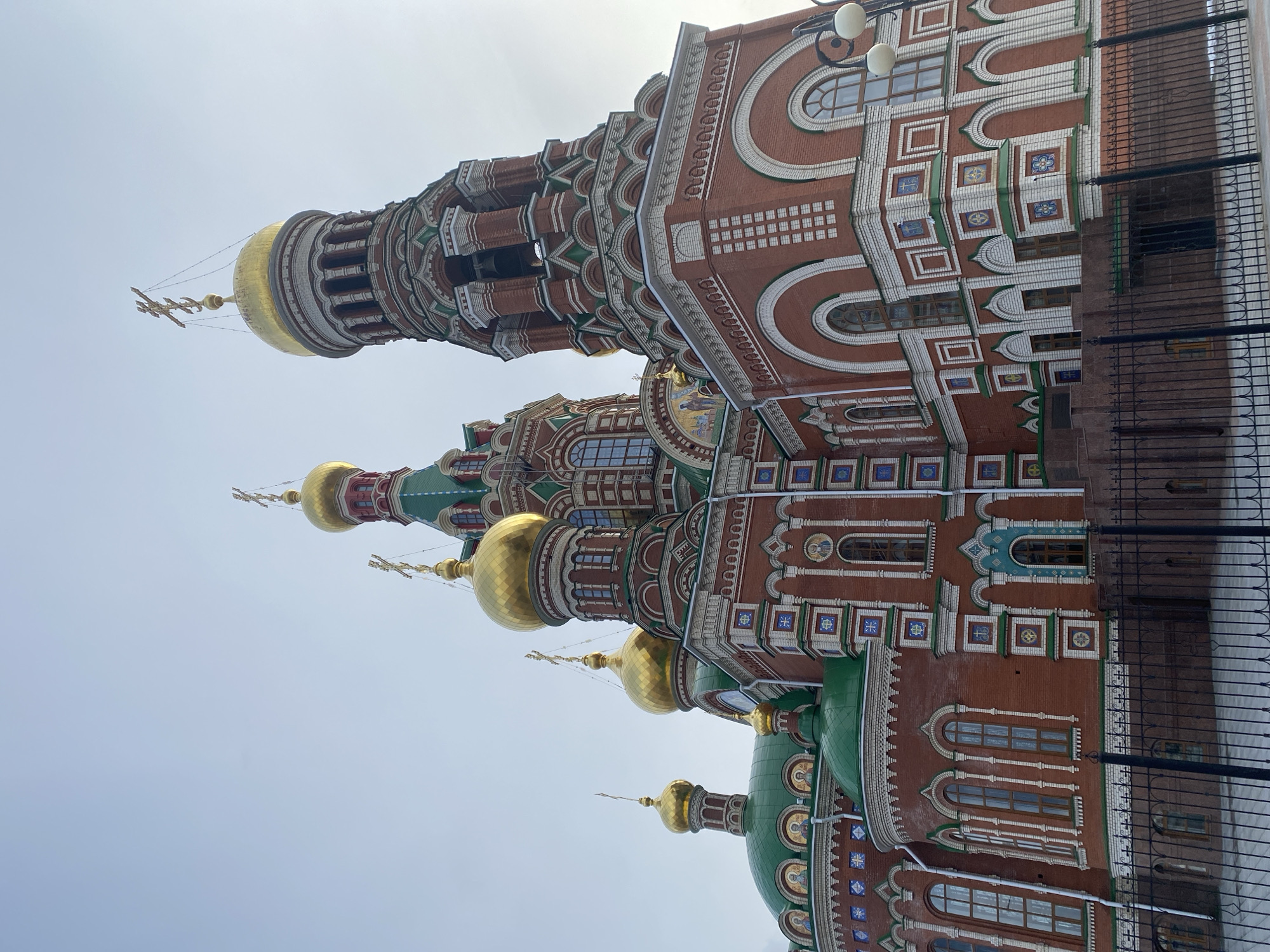 The image displays a grand, ornate Orthodox church building under a bright, overcast sky in Yoshkar-Ola, Russia. The architecture features a predominant reddish-brown brick facade, extensively decorated with white and green trim that creates intricate patterns, arches, and window surrounds.
Multiple golden onion domes are prominent, each topped with a golden Orthodox cross. Some of these crosses are connected by thin wires or chains. Additional domes on the lower sections of the building are green. The structure incorporates tall, cylindrical towers, also adorned with decorative brickwork and featuring arched windows. The facade includes small mosaic panels, some of which appear to depict religious figures or symbols, set within the decorative elements.
The building exhibits a variety of windows, including numerous arched windows and smaller rectangular ones. Lanterns or streetlights are mounted on the upper exterior walls. A black metal fence, comprising vertical bars and decorative scrollwork, runs along the lower right portion of the image, partially bordering the church grounds.
The sky is a uniform, light grey-blue, indicating a bright but overcast daytime. No people or specific activities are visible, allowing the focus to remain entirely on the monumental architecture. The scene captures the static grandeur of the church, characteristic of Russian Orthodox design. There is no discernible text visible in the image.