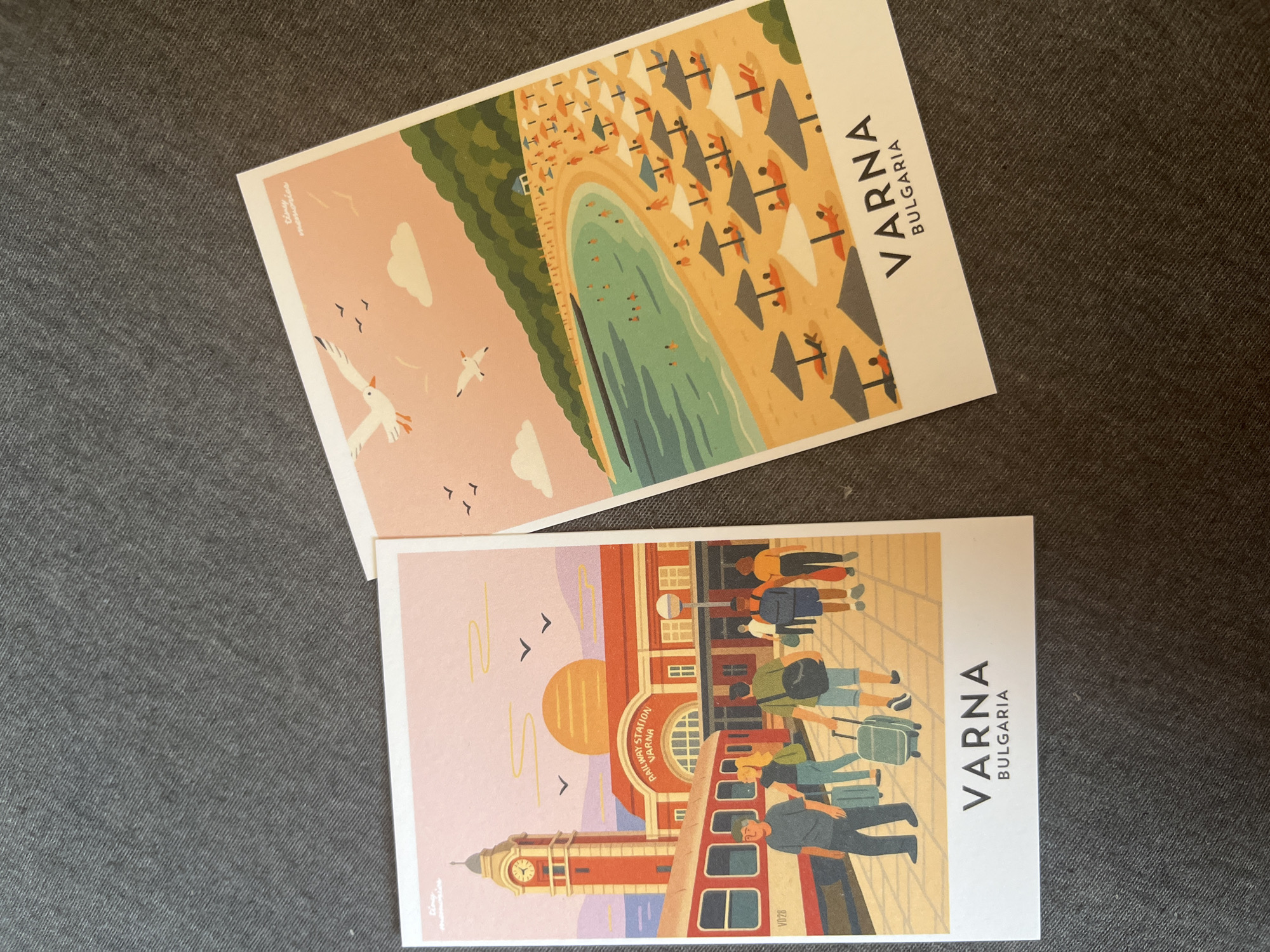 Two postcards are laid on a gray textured surface. The left postcard depicts a stylized illustration of a train station with a red and white train at the platform. A clock tower with a red brick facade stands tall next to the station building, which is also red and features the text "RAILWAY STATION VARNA". Several people are shown walking on the platform with luggage. The right postcard shows a vibrant illustration of a beach scene. People are sunbathing under umbrellas on a sandy beach, and others are swimming in the turquoise sea. Green hills rise behind the beach. Both postcards have text at the bottom. The left postcard reads "VARNA BULGARIA" and the right postcard has "VARNA BULGARIA" in larger font. The city of Montana, Bulgaria is not depicted in this image.