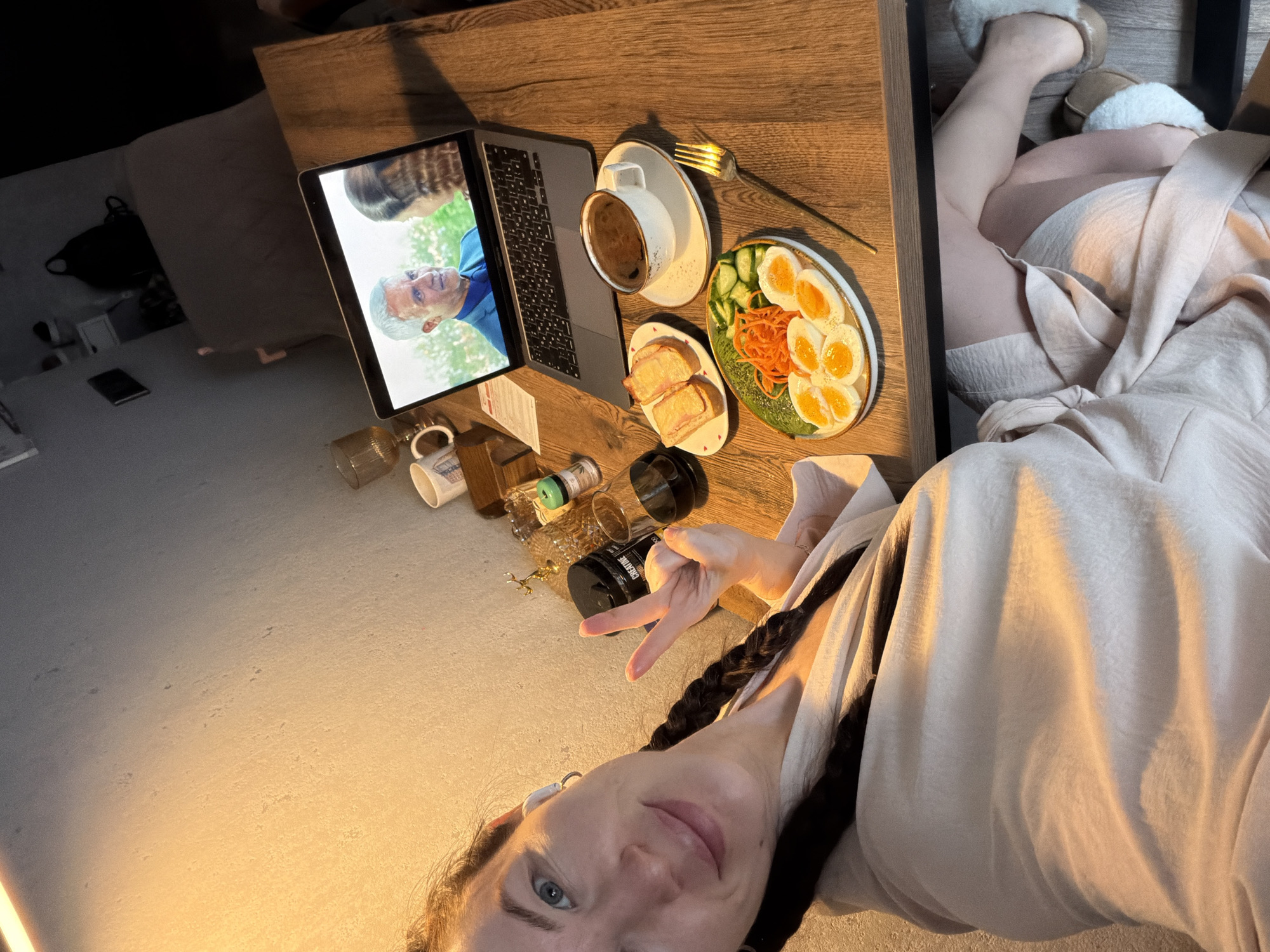 A female subject is captured in a selfie-style portrait, seated at a light-colored wooden table indoors, within what is identified as a room in Perm, Russia. The subject, with dark braided hair falling over her shoulders and a white earbud in her left ear, faces the camera. She is wearing a light-colored robe or loungewear. Her right hand is raised, displaying a peace sign (V-gesture).
On the table in front of her, a meal is arranged. A white plate with a green rim contains several halves of hard-boiled eggs, shredded carrots, and sliced cucumbers. A dark-colored fork rests beside this plate. To its right, a white cup with a brown rim, filled with coffee, sits on a matching saucer. A smaller white plate with red heart decals holds two pieces of toast topped with a pinkish spread.
Further back on the table, a silver laptop is open, displaying a video scene featuring a smiling male character with gray hair wearing a blue shirt, interacting with a female character seen from the back. Adjacent to the laptop, a clear glass contains a dark liquid. A large black container labeled "CREATINE" is partially visible. Additional items on the table include a small gold-colored decorative object, another clear ribbed glass, and a light-colored mug with blue patterns.
The subject's lower body is partially visible beneath the table, with her legs crossed and wearing light-colored, possibly fur-lined, slippers. The background wall is light-colored and textured, illuminated by a warm light source from the upper left. The overall room appears dimly lit, with faint details in the far background suggesting a bed with a light-colored headboard, a dark backpack, and a framed item on the wall.