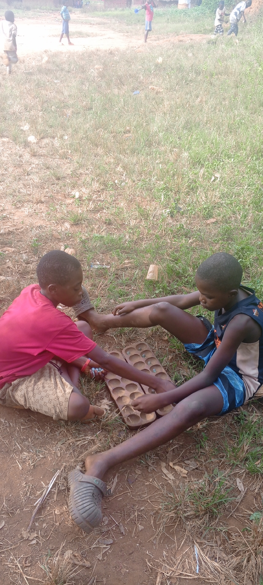 Two young boys are seated on the ground in an open, grassy area, likely in Ngalula, Democratic Republic of the Congo. They are playing a game on a wooden board with multiple indentations. One boy wears a red shirt and patterned shorts, while the other has on a dark tank top and blue shorts. Both are barefoot. In the background, other children appear to be playing. The ground is a mix of dirt and grass, with dry leaves and twigs scattered around.