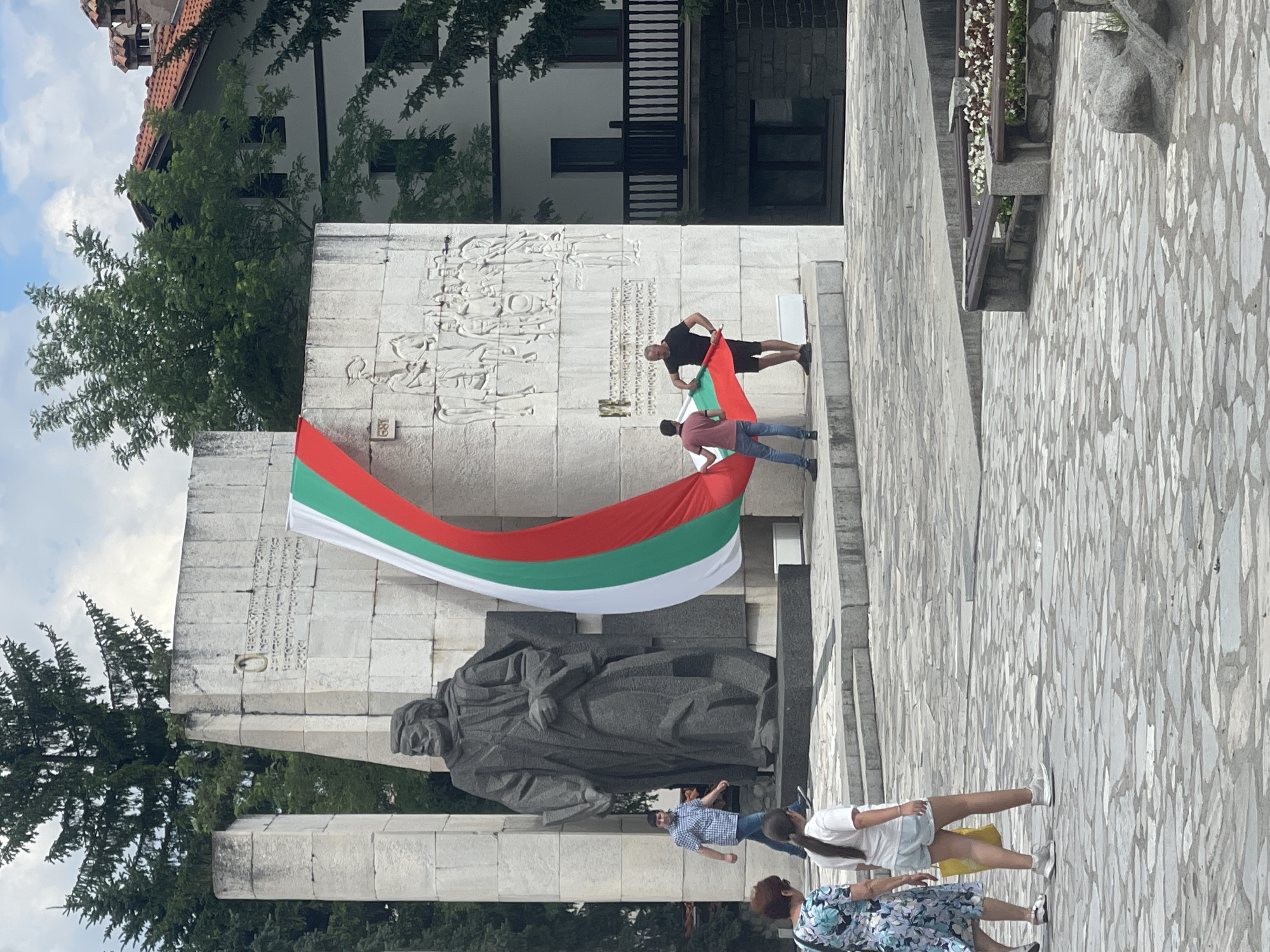 In the city of Unknown, Unknown, two men are seen holding a large Bulgarian flag. The flag is draped over a stone monument, which features a large statue of a man on the left and relief carvings on the right. One man, wearing a pink shirt and blue jeans, is positioned on the left side of the flag, while the other man, in a black t-shirt and shorts, is on the right. In the background, a woman in a floral dress and a young girl in a white t-shirt and light shorts are walking away from the monument. The ground is paved with irregular stone slabs. Trees and a building with a red tiled roof are visible behind the monument. The sky is partly cloudy.