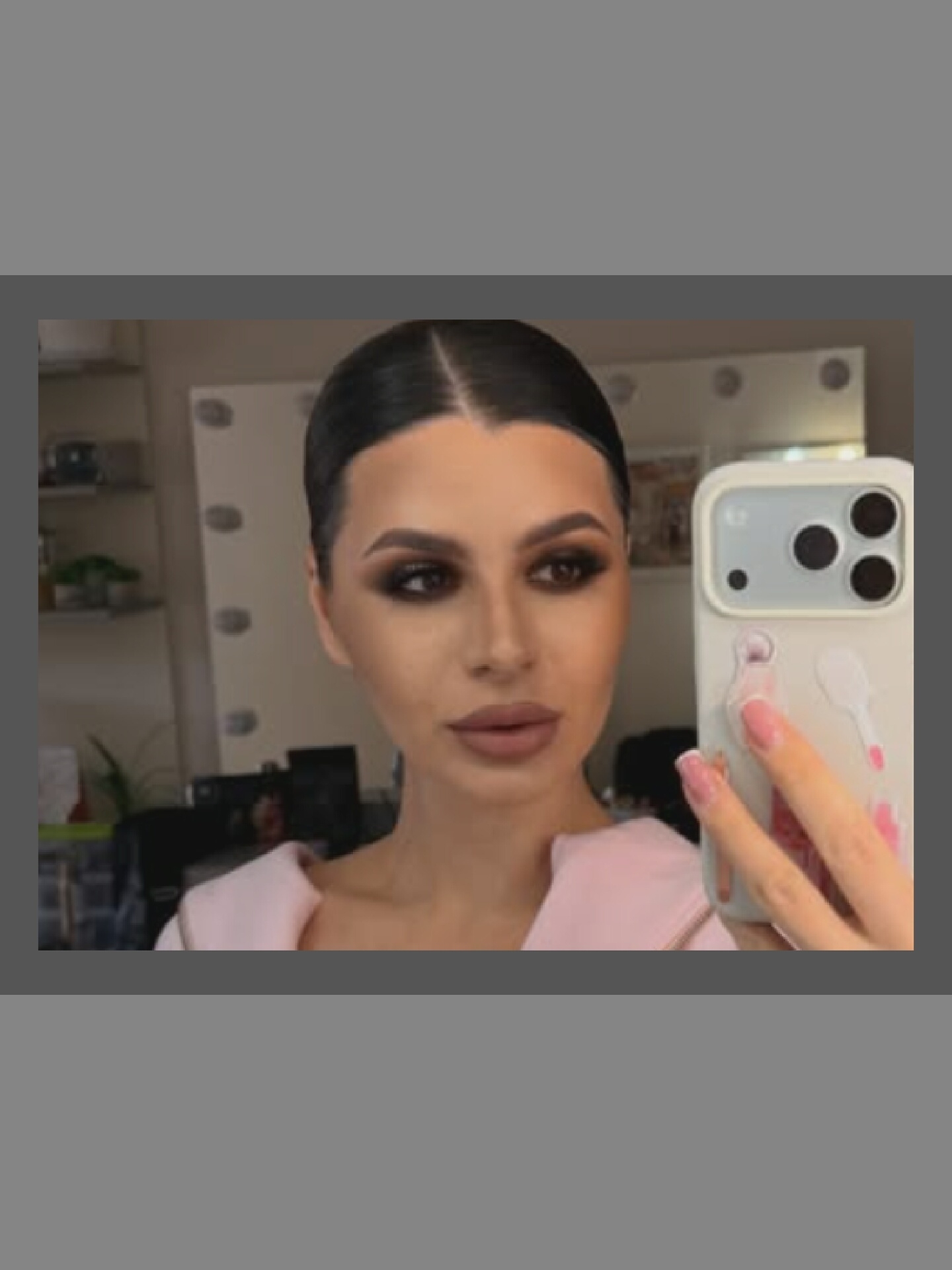 A woman with dark hair pulled back and dramatic eye makeup takes a selfie in a well-lit room with mirrors. She is wearing a pink top. Her right hand holds a white phone with a decorative case and a camera. The background shows shelves with various items and lighting fixtures. The room appears to be a dressing room or studio. The location is Unknown, Unknown.