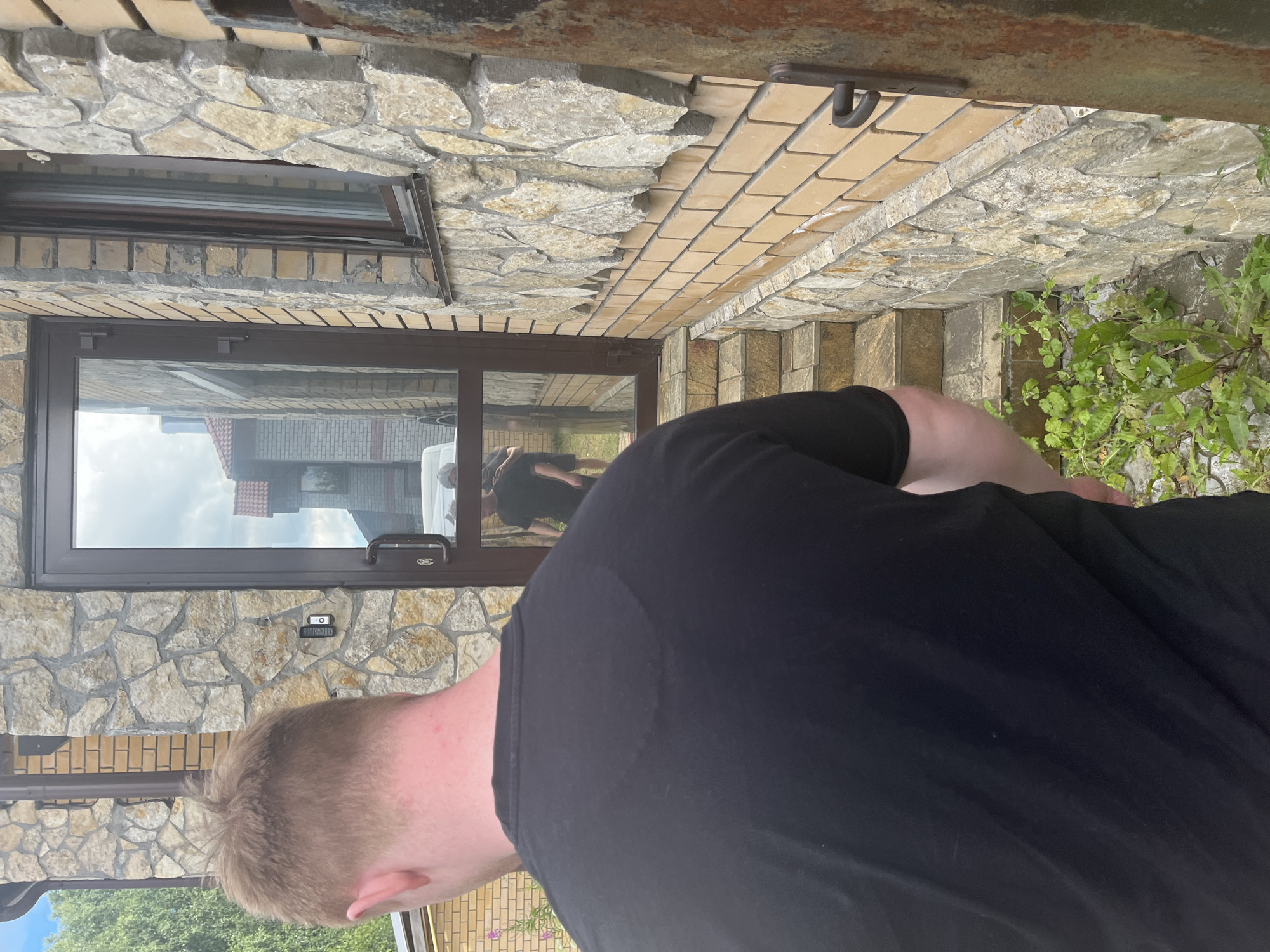 A person wearing a black t-shirt is standing with their back to the camera, facing a glass door. Their hair is short and light brown. The building is made of stone and brick. In the reflection of the glass door, another person in a black t-shirt and shorts is visible, along with a white van. There are steps leading up to the door. Green plants are growing on the ground to the right of the steps. The scene appears to be outdoors, possibly in front of a residential building. The city is specified as Orekhovka, Russia.