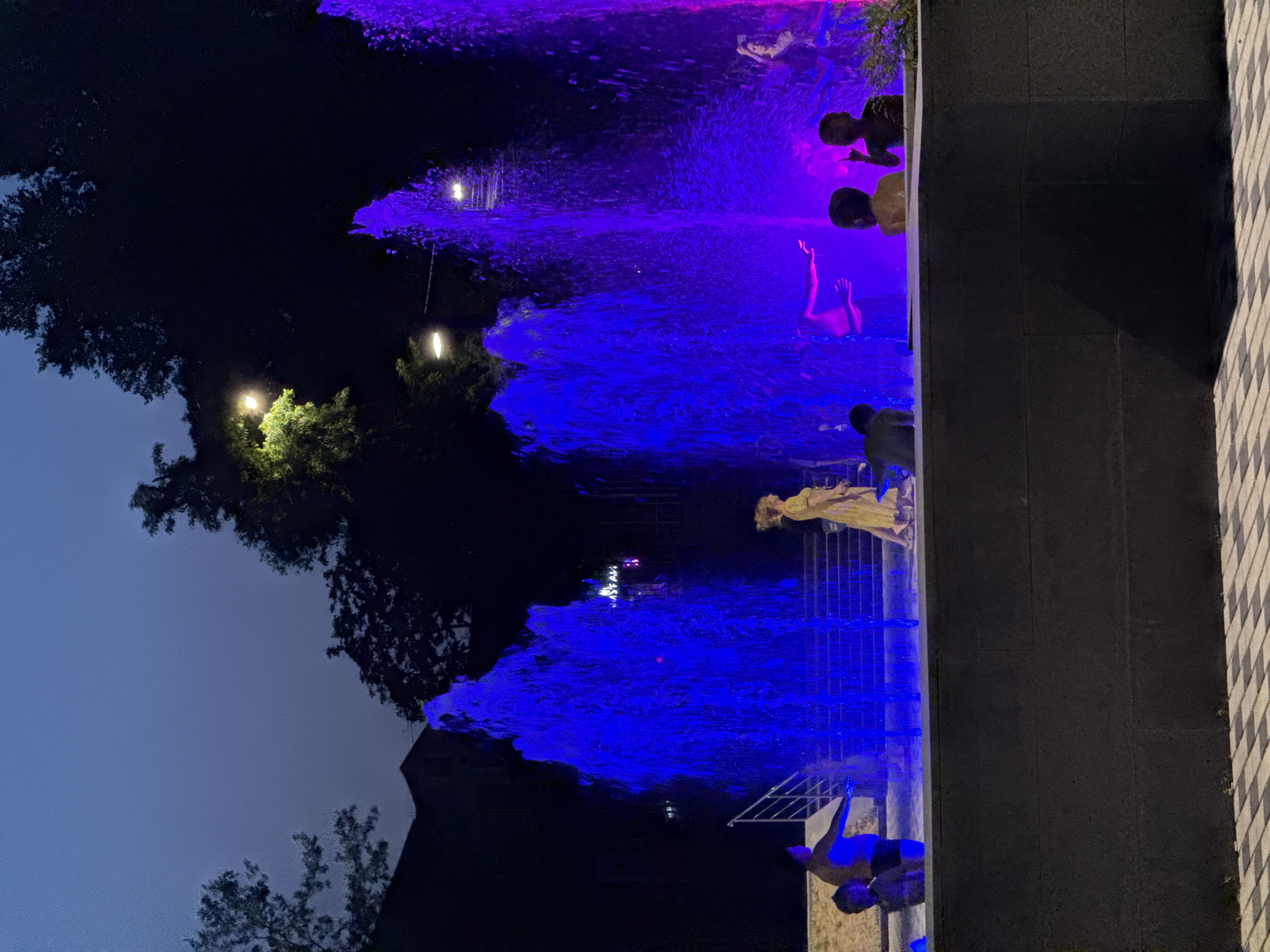 This image depicts a public fountain at night, illuminated by vibrant blue and purple lights. Several individuals, including adults and children, are present near the fountain. Some are standing and observing, while others appear to be interacting with the water. The background includes trees, buildings, and streetlights, suggesting an urban setting. The presence of a sign in Cyrillic text, partially visible in the upper right portion of the image behind the purple fountain, suggests the location is likely in Russia. Given the architectural style and the context of public fountains, this scene could be in Rostov-on-Don, Russia.
