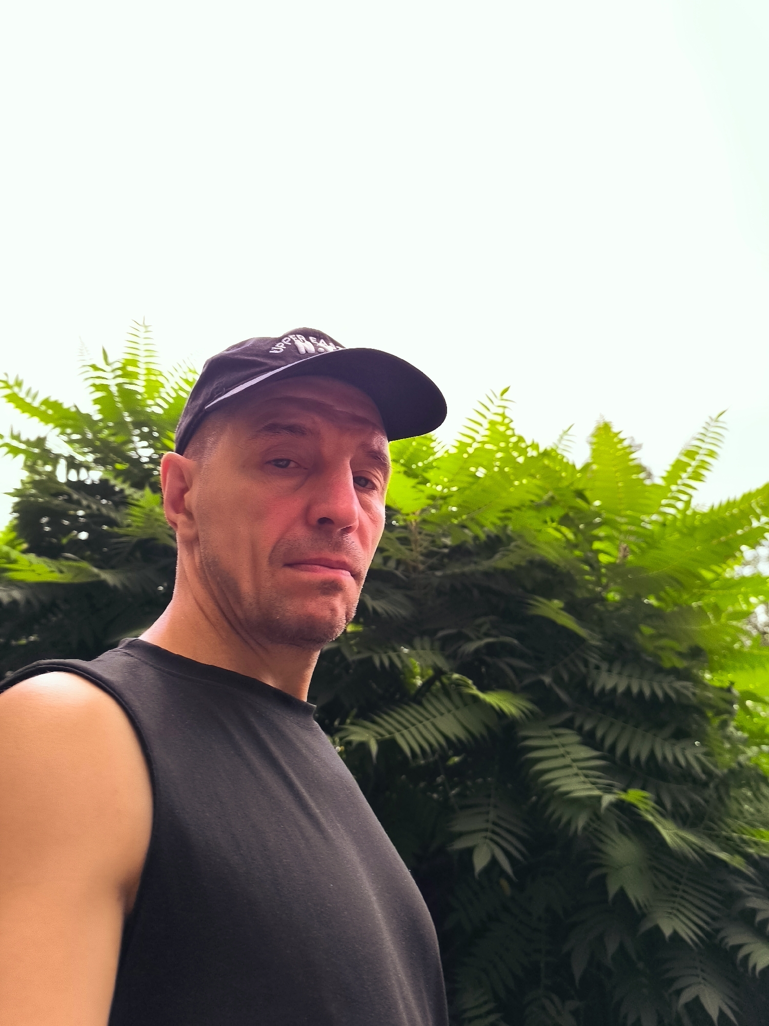 A man wearing a black sleeveless shirt and a black baseball cap is standing outdoors, looking at the camera. He has a neutral expression on his face and is positioned on the left side of the frame. Behind him, lush green foliage from a tree is visible, filling the right side of the image. The background is a bright, overcast sky. The image appears to have been taken in Almaty, Kazakhstan, possibly in a park or residential area with abundant greenery.