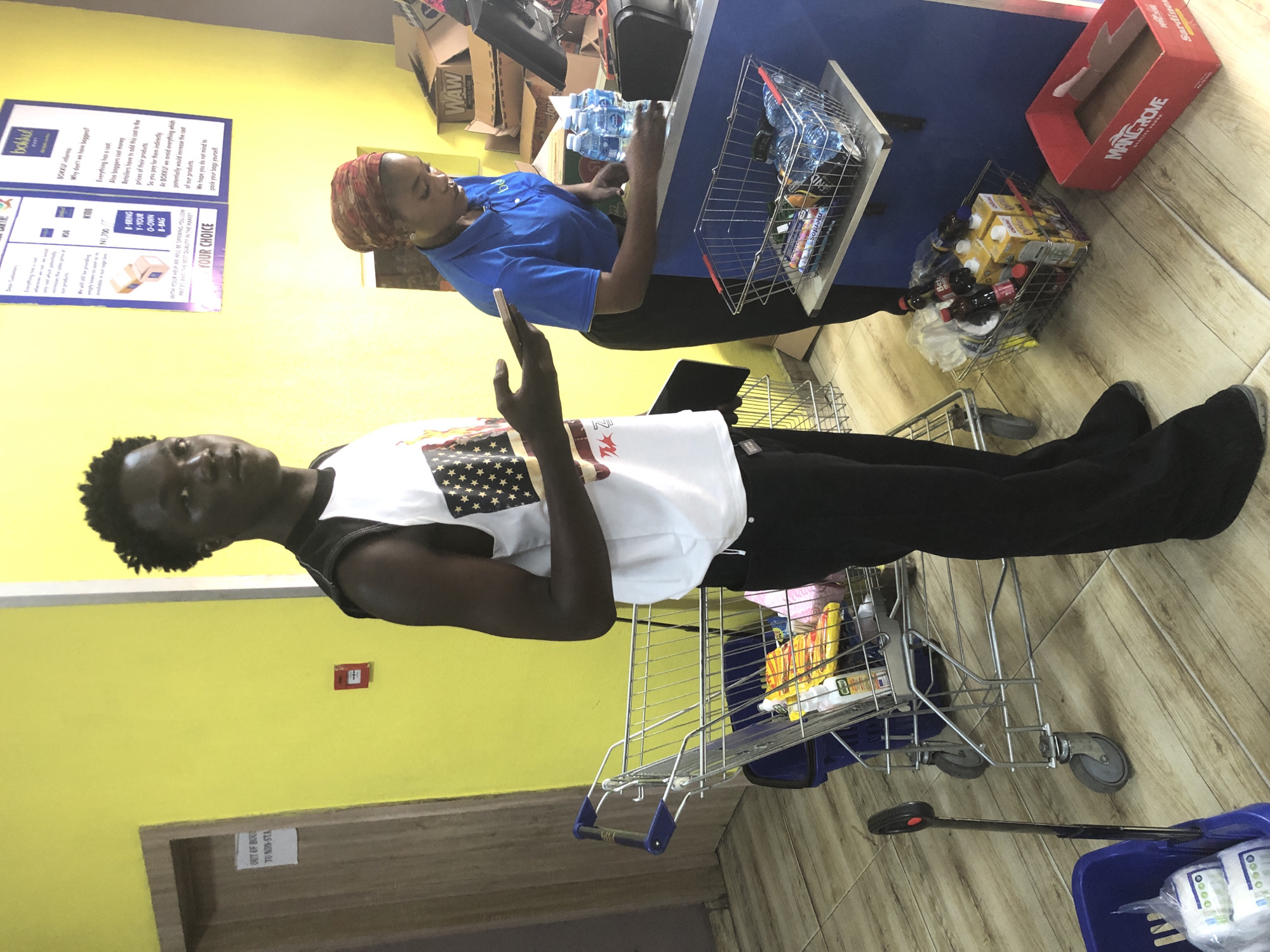 In Ado, Nigeria, inside a brightly lit store with yellow walls and light-colored tiled flooring, a young Black male stands near a shopping cart. He is wearing a white sleeveless shirt with a black trim, featuring an American flag print on the front, and black trousers. He is holding a smartphone in his left hand and a tablet in his right. Beside him, a Black female, possibly an employee, is positioned behind a blue counter. She is wearing a blue polo shirt and a headscarf, and she is reaching for bottles of water on the counter. A shopping basket containing various items, including bottled water and snacks, is placed on a shelf below the counter. Another shopping cart is visible to the left of the male, containing similar items. A red box with "MANGROVE" printed on it is on the floor to the right. Signs are posted on the yellow wall behind the counter.