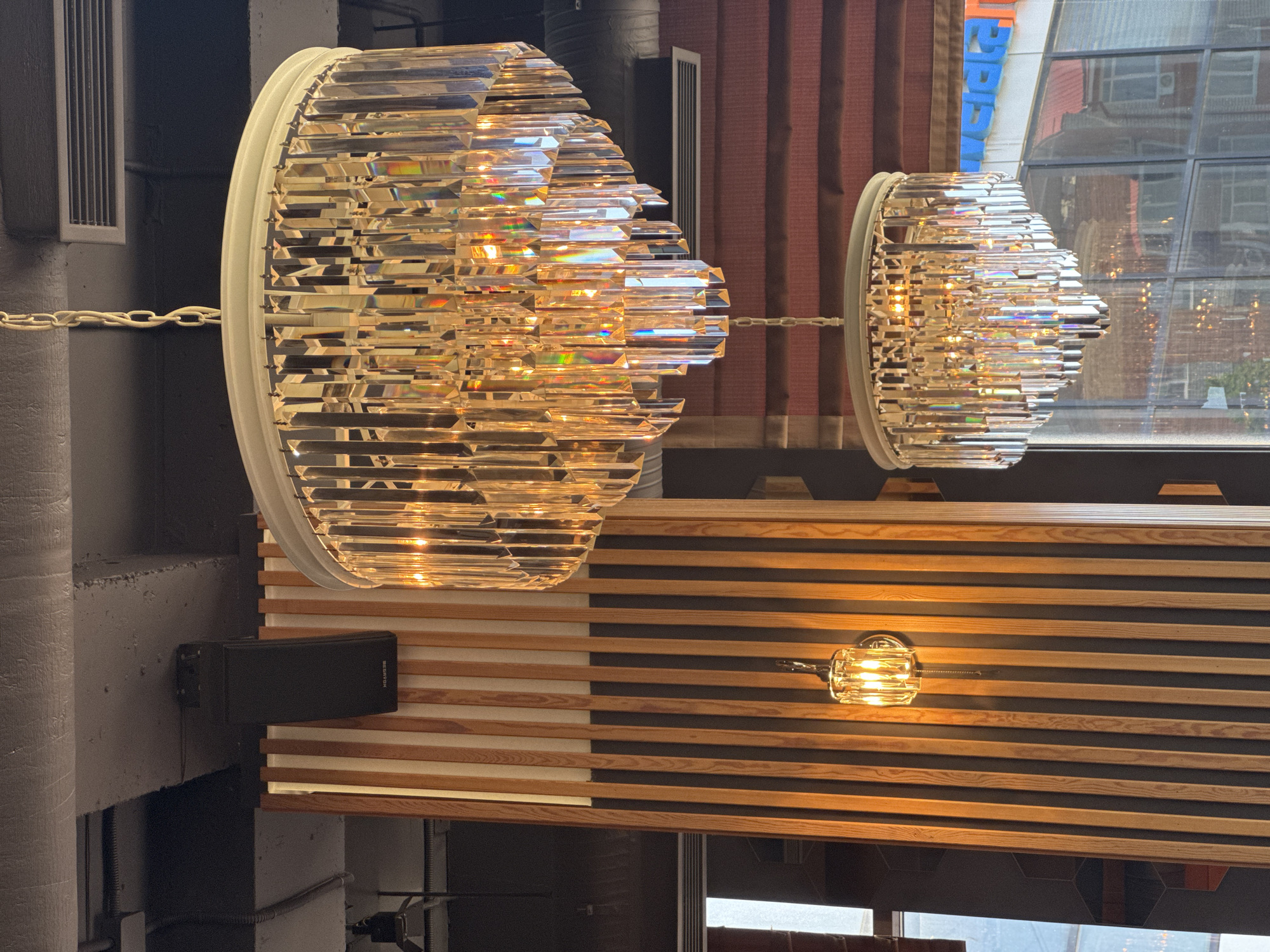 The image shows two large, faceted crystal chandeliers hanging from a ceiling. The chandeliers are illuminated, casting a warm glow and reflecting light. They are suspended by white chains. Below the chandeliers, there is a decorative wall paneling made of horizontal wooden slats alternating with dark panels. A small, clear glass light fixture with a faceted glass shade is mounted on this paneling. To the left of the paneling, a black speaker is mounted on a concrete pillar.
The scene appears to be indoors, possibly a restaurant or a commercial space, given the decorative elements and the presence of speakers. The lighting suggests it could be evening or a dimly lit interior space. Through a large window in the background, a cityscape is visible, with buildings and reflections suggesting it is daylight outside. The overall impression is one of modern, sophisticated interior design with a focus on lighting and materials. No people are visible in the image. There is no visible text.