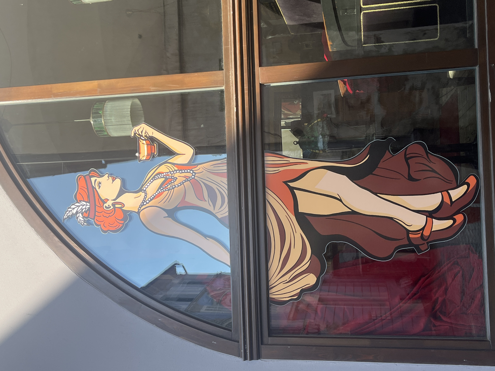 This media file shows a large graphic decal of a stylized woman in a vintage Art Deco style, displayed on a storefront window. The decal is divided into two sections, covering two adjacent panes of glass. The woman is depicted in profile, with reddish-orange hair styled in a bob and a feathered headband. She is wearing a long, flowing dress with abstract patterns in shades of beige, orange, and brown, and a beaded necklace. In her right hand, she holds a small, orange object that appears to be a compact or lipstick case, and she is looking at it. Her legs are crossed, and she is wearing brown heels with an orange strap.
The decal is applied to a window that appears to be part of a shop or cafe. The background of the decal in the left pane is a clear blue sky, suggesting an outdoor scene or an aspirational depiction. Reflections are visible in the glass, showing a dimly lit interior with some furniture and fabric, possibly a red velvet cloth draped in the background of the right pane. The wooden frame of the window is visible, with a prominent vertical divider between the two panes.
The overall impression is one of elegance and a bygone era, likely representing a brand or theme associated with vintage aesthetics, perhaps a salon, a boutique, or a cafe. There is no visible text within the image. The lighting suggests it might be daytime, as sunlight is casting shadows on the exterior wall to the left. The reflections indicate an indoor setting behind the window, but the activity depicted by the decal is static.