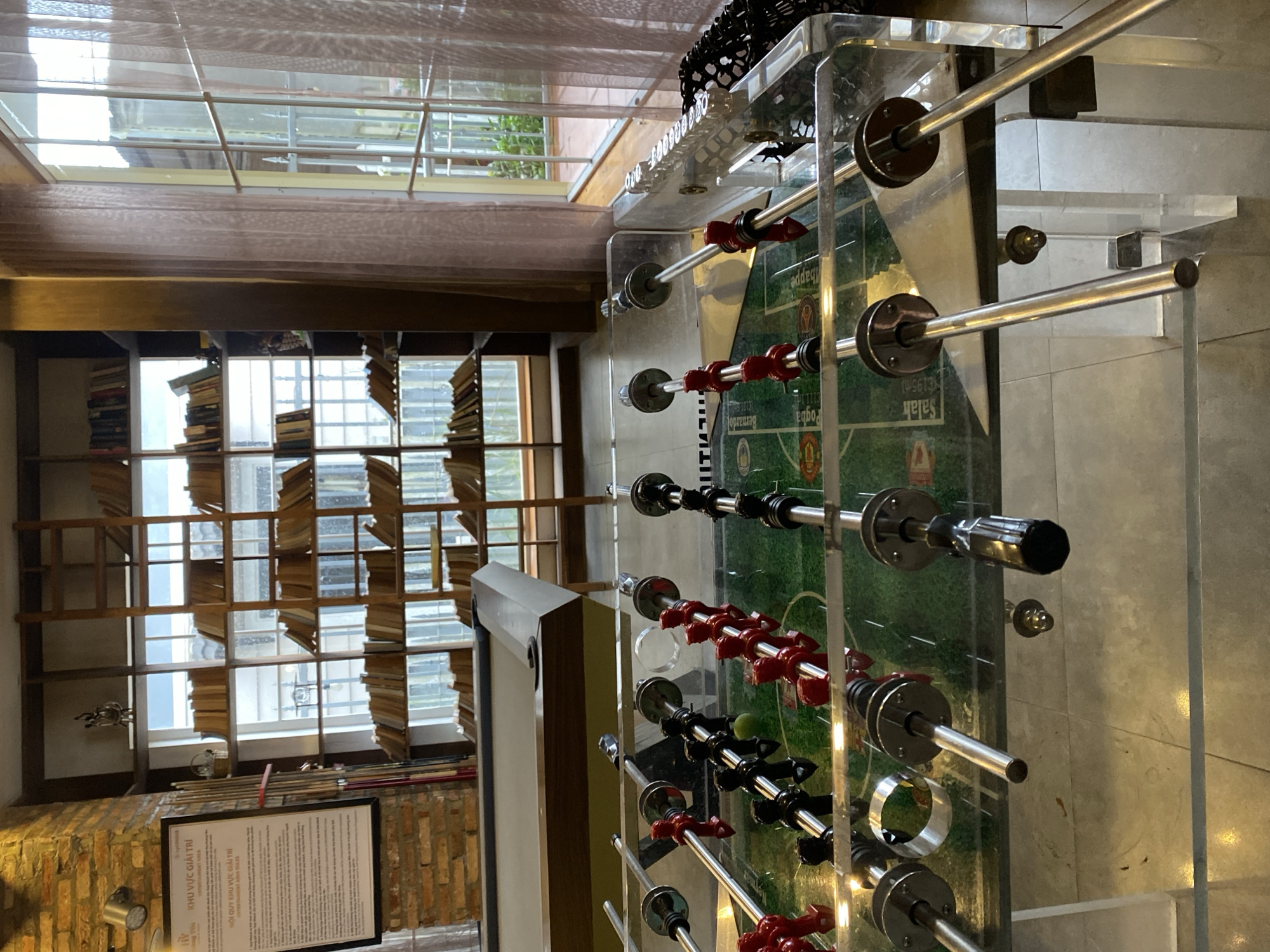 The image shows a foosball table in the foreground, with a pool table and bookshelves in the background. The foosball table is clear acrylic and has red and black players. The pool table is gray with a wooden edge. The bookshelves are filled with books and are very tall, reaching almost to the ceiling. There are also large windows with sheer curtains on the right side of the image. The room appears to be well-lit and has a modern, stylish feel. The text on the sign in the background suggests that this location is in Da Lat, Vietnam, specifically an "ENTERTAINMENT AREA."