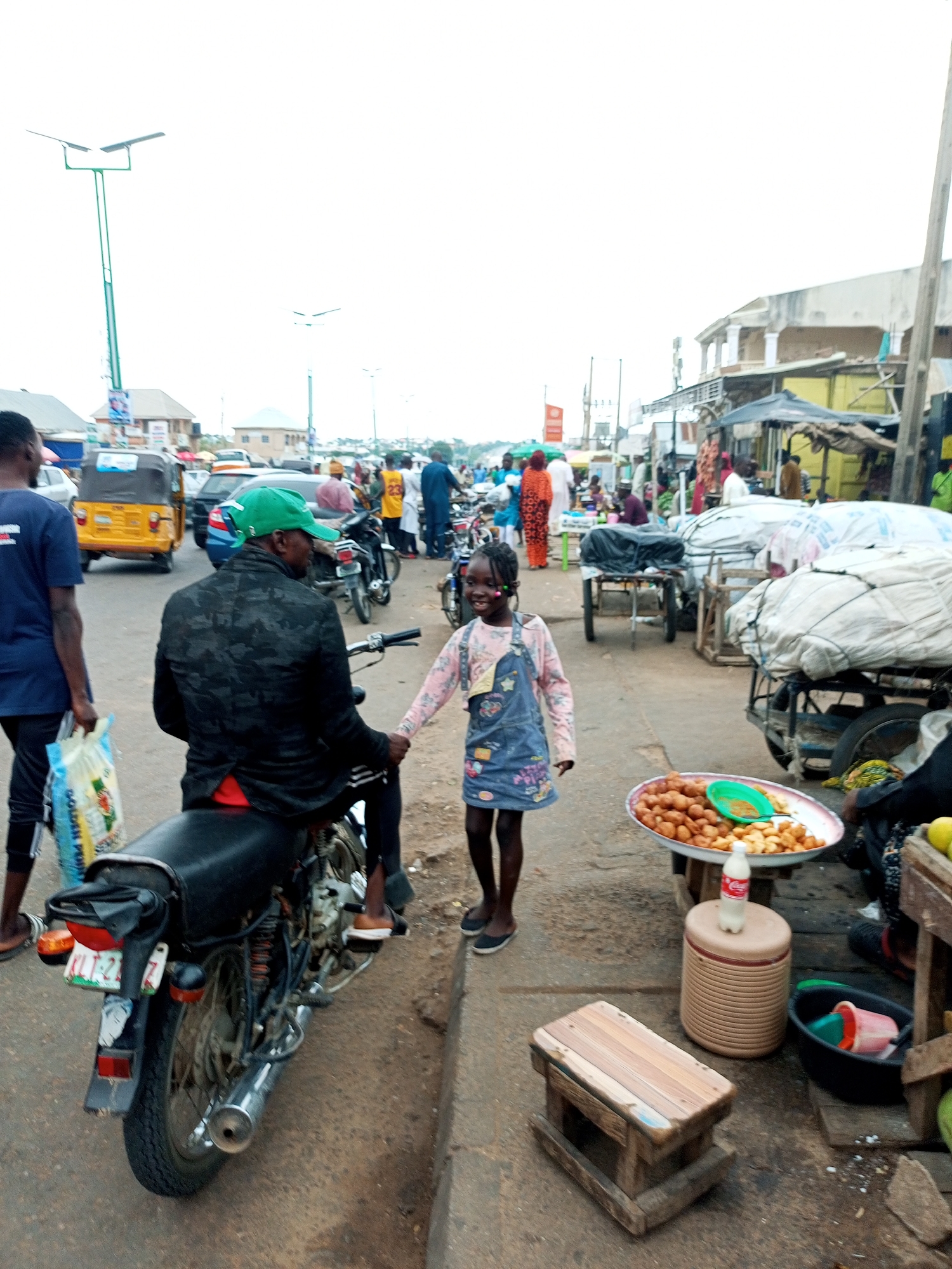The image depicts a street scene in what appears to be a marketplace. In the foreground, a man wearing a camouflage jacket and a green cap is seated on a motorcycle, facing away from the camera. Beside him, a young girl in a denim overall and a pink long-sleeved shirt is smiling and holding his hand. She is standing next to a table displaying a platter of fried snacks. Behind them, a yellow tricycle is visible, along with other motorcycles and people in the background. Buildings line the street, and there are various market stalls with goods covered by tarps. The overall environment suggests an outdoor commercial area with moderate pedestrian and vehicular activity. The sky is overcast.