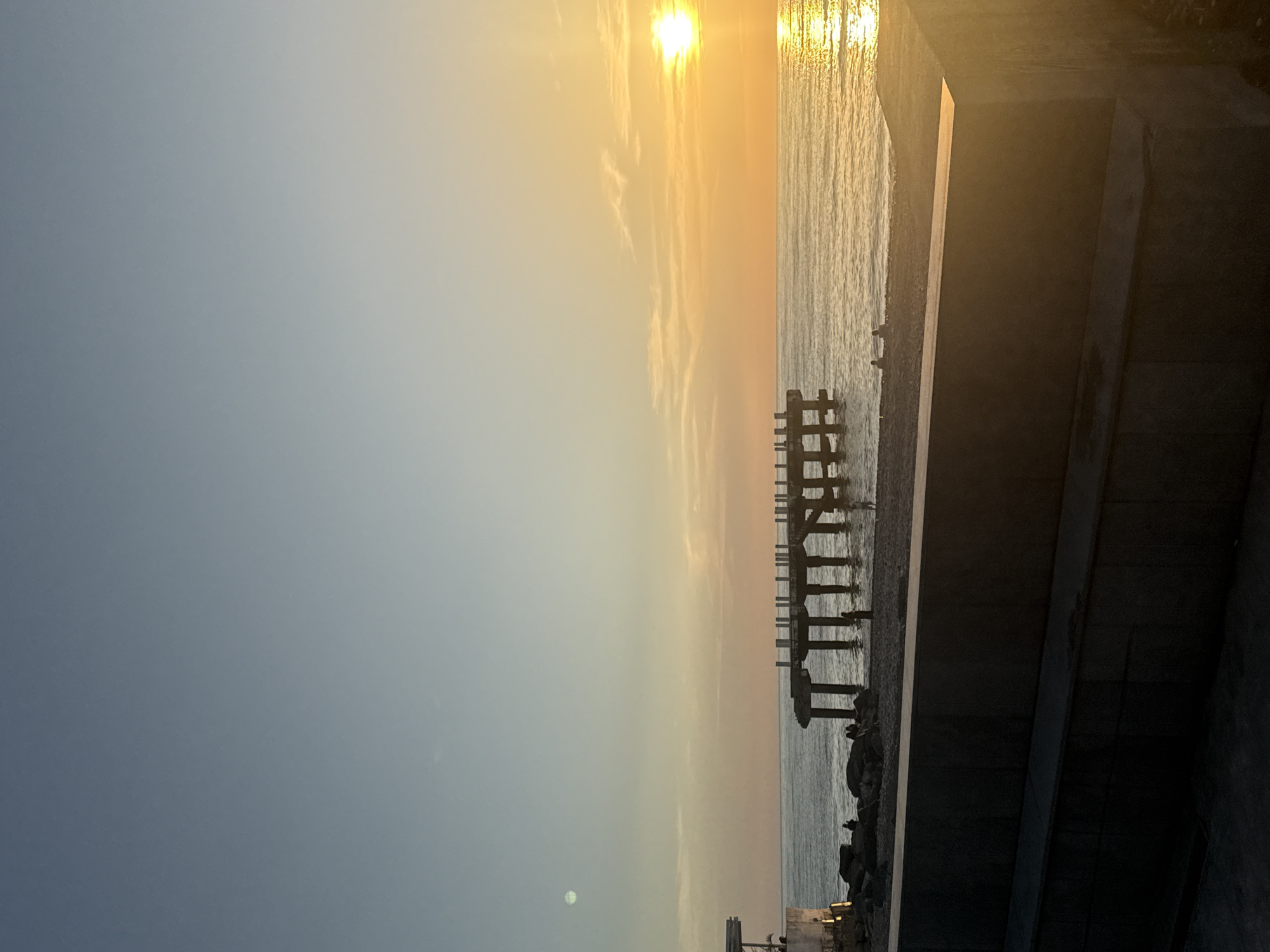 The image depicts a seascape at sunset. The sun, a bright white disc, is positioned over the water, reflecting light onto the sea's surface. To the right of the frame, a dark concrete structure is visible, partially obscuring the view of the water. A derelict pier stretches into the sea, its supporting pillars and beams silhouetted against the bright sky. The sky transitions from a light blue on the left to an orange hue towards the horizon.
Based on the derelict pier structure and coastal features, this appears to be a view of the Black Sea in Batumi, Georgia.