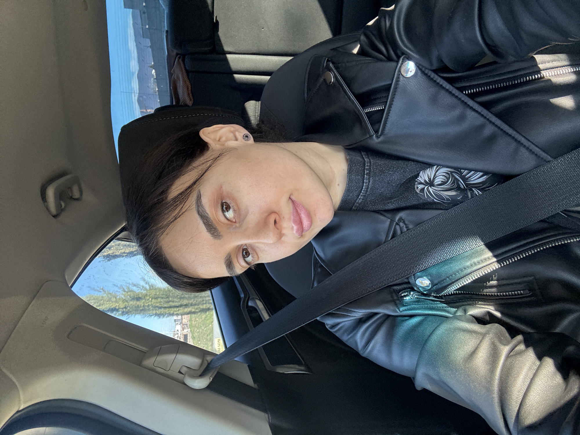 A woman is in a car, wearing a black leather jacket and a seatbelt. The background visible through the car window shows a landscape, including trees and buildings, suggesting the location is in Tbilisi, Georgia.
