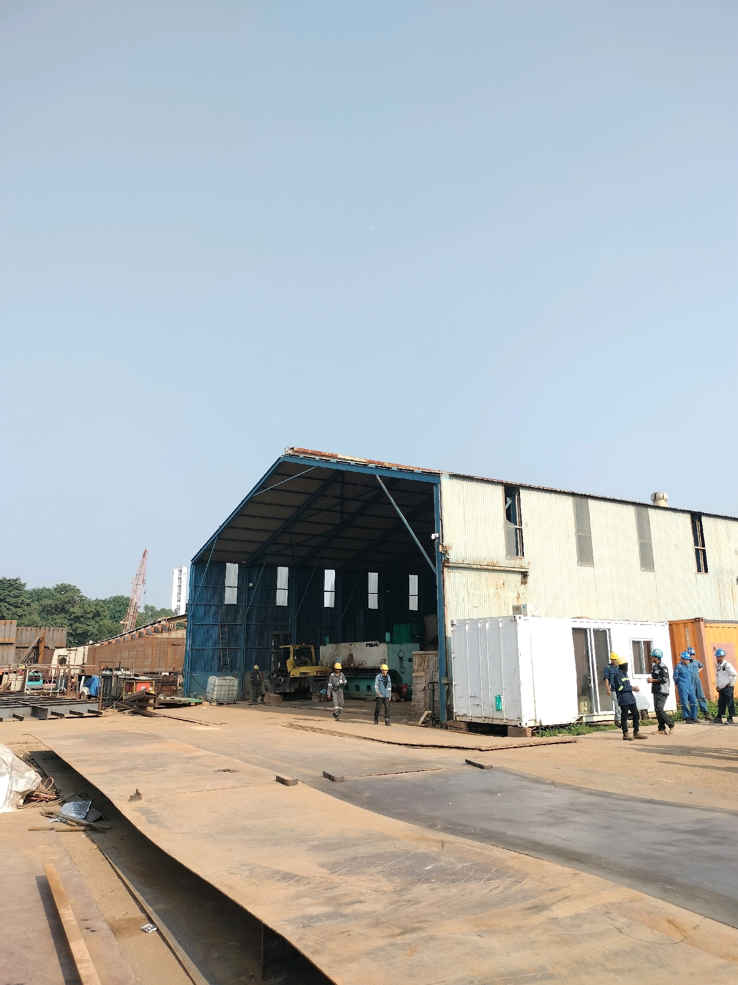 The image shows an outdoor industrial scene with a large, open-sided warehouse in the center, likely a fabrication yard or shipyard. The warehouse has a corrugated metal roof and siding, with open bays. Inside the warehouse, a yellow forklift is visible, along with some machinery. Outside the warehouse, several people in work clothes and hard hats are gathered. To the right of the warehouse are several portable container-like structures, one white and one orange. The foreground consists of large, flat metal plates laid out on the ground, suggesting ongoing construction or fabrication. In the background, a crane is visible, along with more industrial structures and trees. The sky is clear and light blue, indicating daytime. The overall impression is of an active industrial environment. This scene is consistent with a shipyard or industrial complex in Batam, Indonesia, known for its shipbuilding and manufacturing industries.