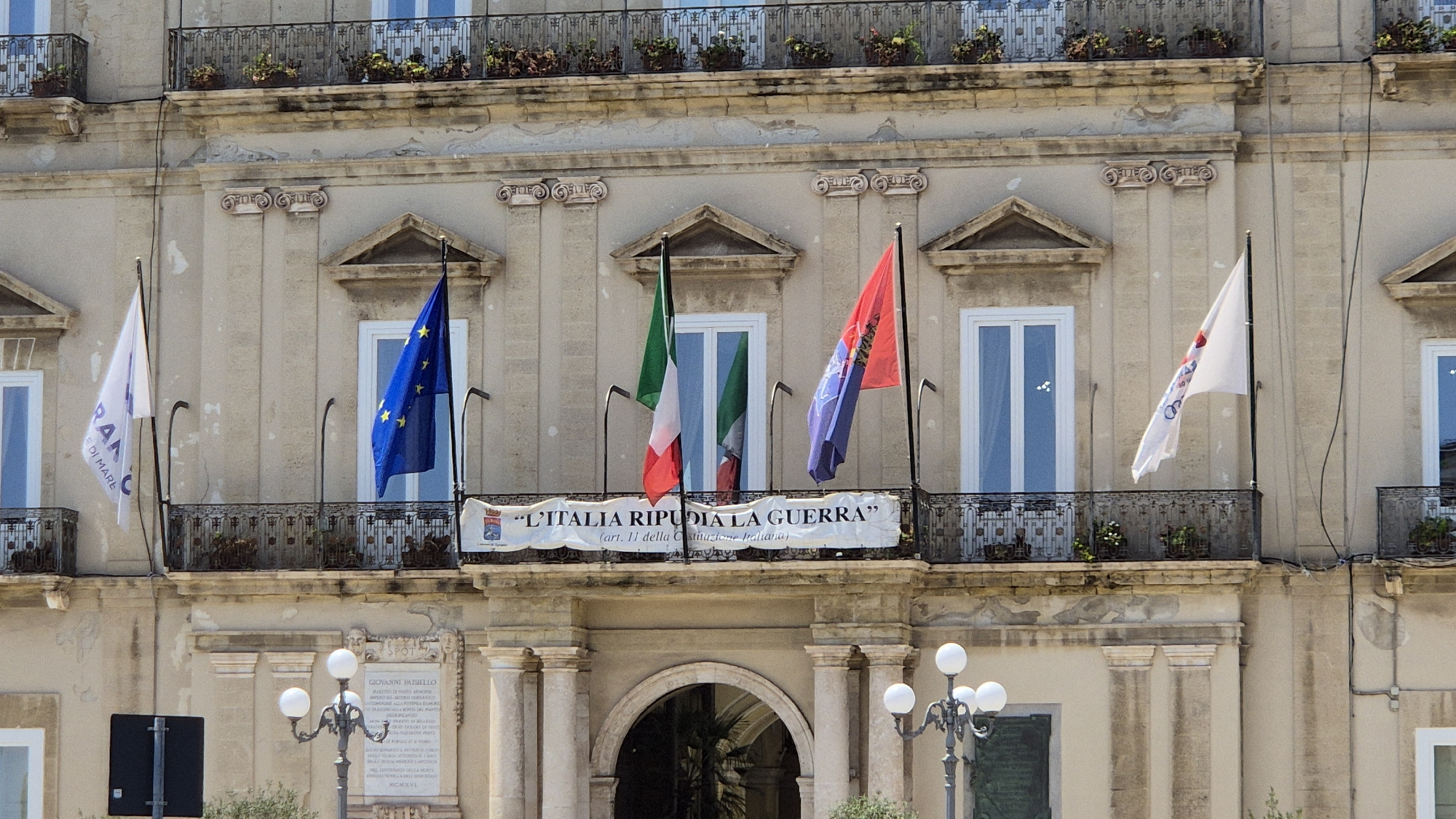 The image shows a facade of a building, likely a public building given the presence of flags and a banner. The building is made of light-colored stone and features balconies with ornate railings on multiple levels. Several flags are displayed on poles extending from the balconies. From left to right, the flags include a white flag with blue lettering that is partially visible, a European Union flag, an Italian flag, and a red and blue flag, possibly a regional or organizational flag. There is also a white flag with red and blue emblems on the right side of the image.
A banner is stretched across a balcony in the center of the facade, displaying text in Italian that reads "L'ITALIA RIPUDIA LA GUERRA" (Italy rejects war), followed by "(art. 11 della Costituzione Italiana)" (article 11 of the Italian Constitution). This suggests the building might be a town hall, government office, or historical site related to Italian law and culture.
Below the balconies, there is an arched entrance leading into the building, with visible greenery and shadows inside. Two decorative street lamps with multiple globes are visible in the foreground, to the left and right of the entrance. A plaque is mounted on the building to the right of the entrance, with Italian text inscribed on it, likely commemorating a historical figure or event. The overall impression is one of a civic or historical commemoration taking place in a public space in Taranto, Italy.