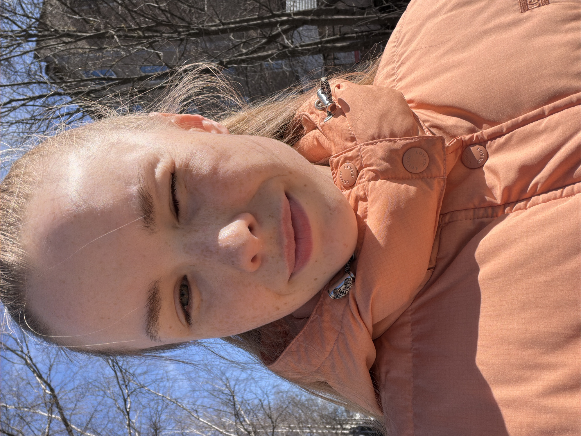 The image is a close-up, slightly tilted portrait of a fair-skinned woman with blonde hair and freckles. She is wearing an orange puffer jacket. Bare trees and a building can be seen in the background. This is likely a winter or early spring scene. I am unable to verify if this image contains the city of Berezniki, Russia.
