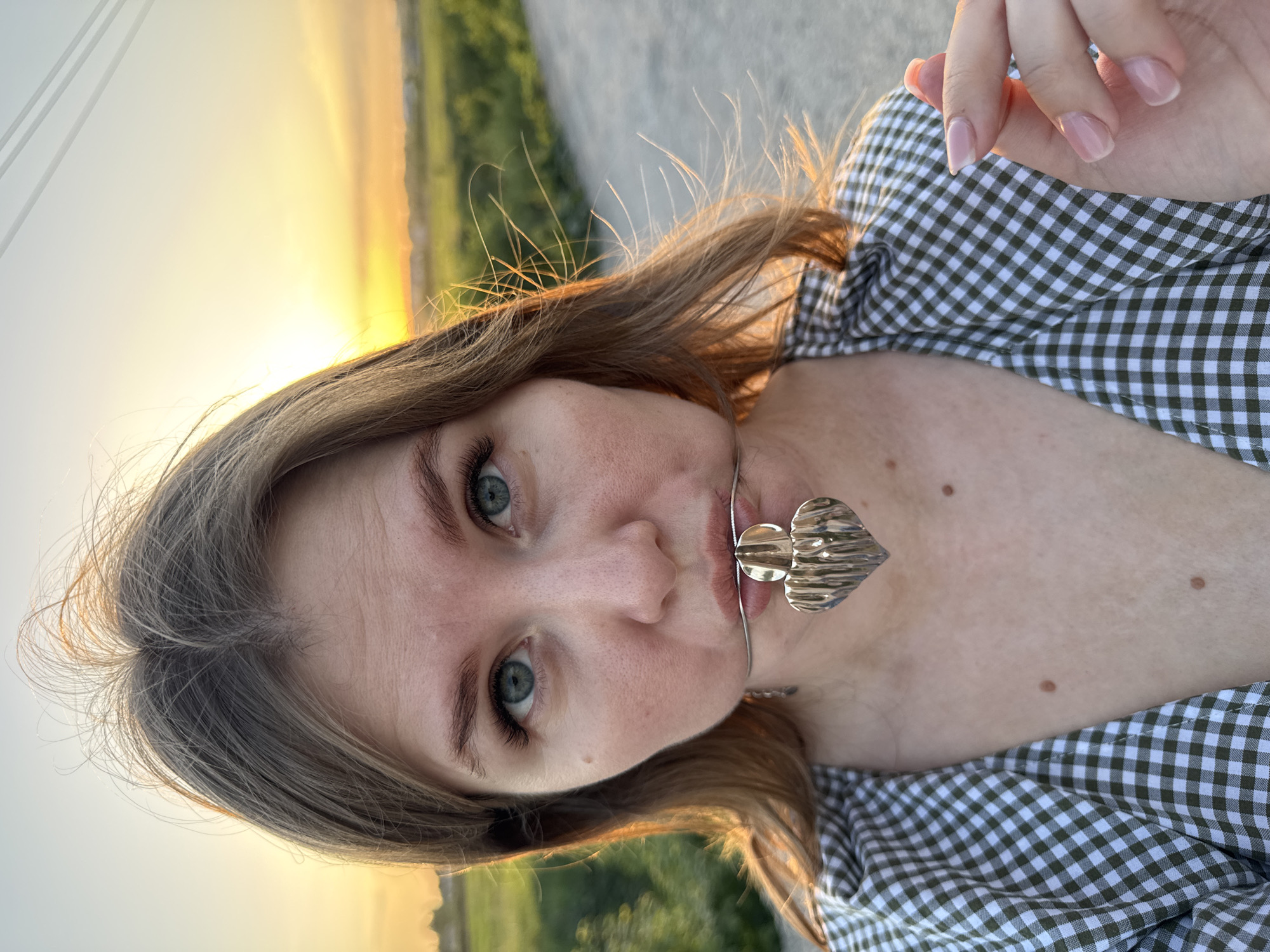 A light-skinned woman with shoulder-length brown hair is centered in the frame. She has blue eyes and light eyebrows. She wears a green and white gingham shirt. A silver-colored choker necklace with a heart pendant is around her neck. The background features a sunset over a green field and a road. The picture was taken outdoors in daylight. Location: Горный, Russia.