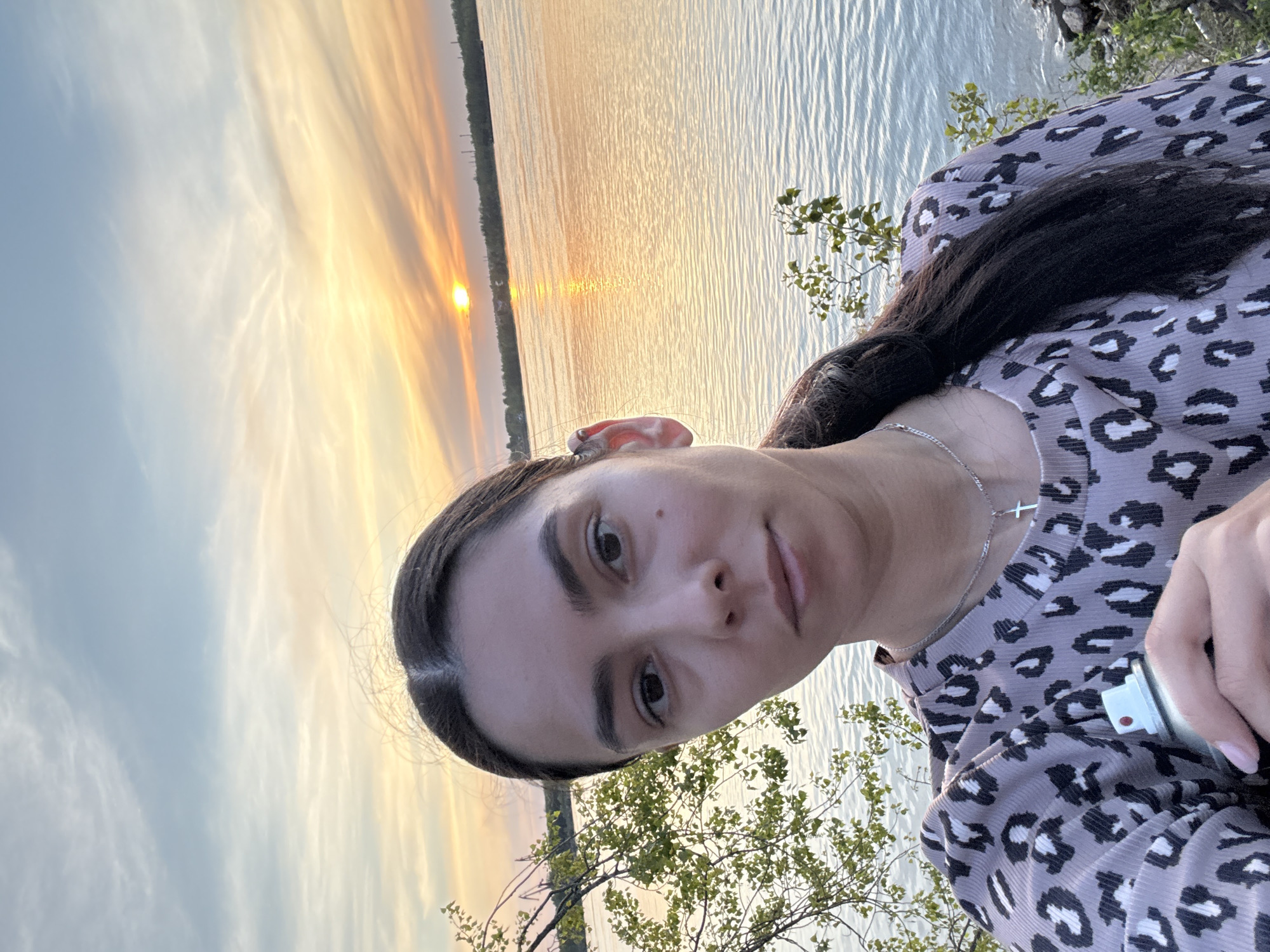 A woman is standing outdoors with a body of water and a sunset in the background. The woman has dark hair pulled back, fair skin, and is wearing a patterned shirt and a silver necklace with a cross pendant. She is holding a cylindrical object in her right hand. The water reflects the sunset colors, and trees are visible along the shoreline. The sky displays a mix of yellow, orange, and blue hues. Location is unknown.