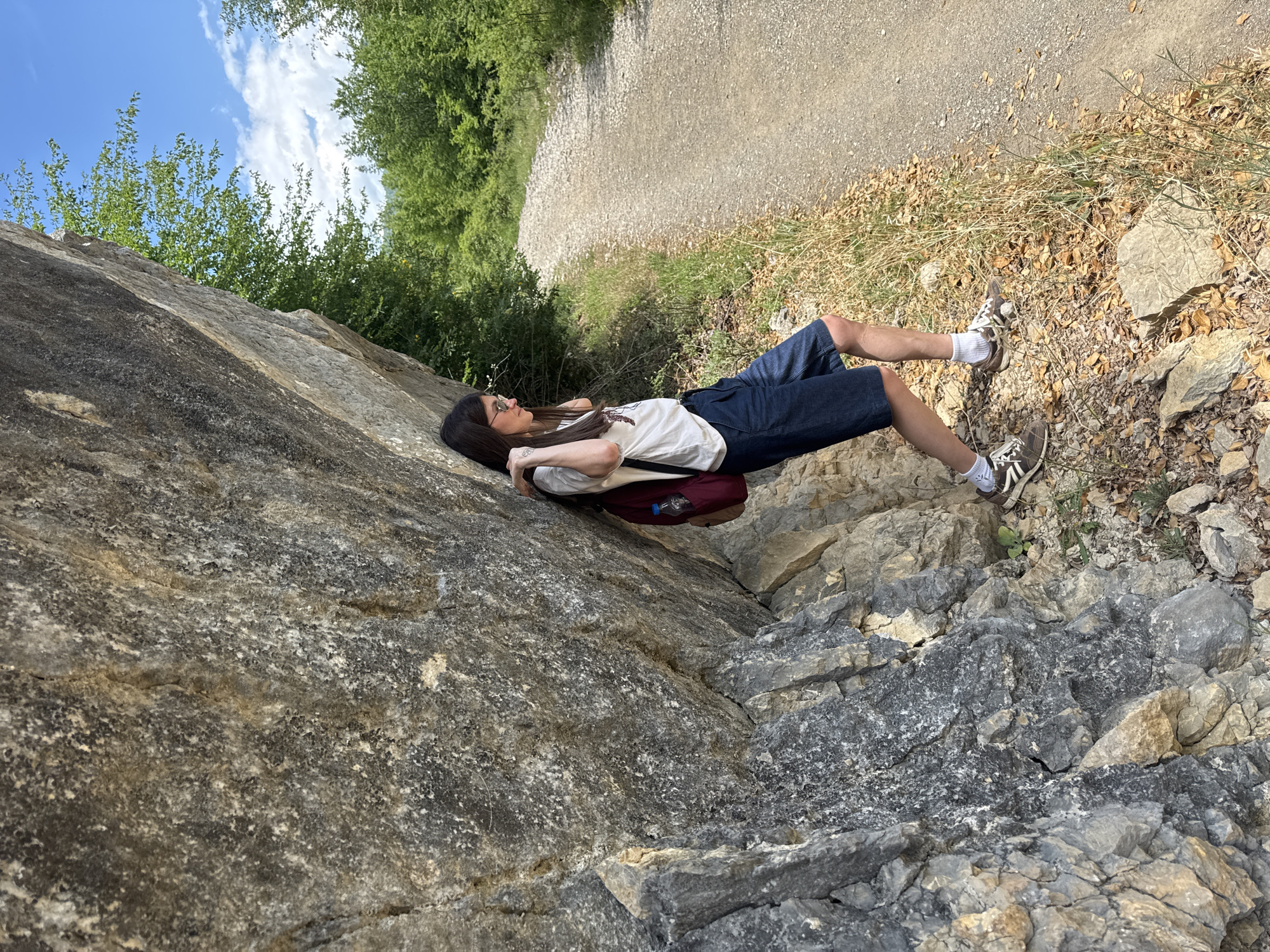 A woman is lying on a rock face adjacent to a dirt road and grassy area, seemingly in Mrtvo Duboko, Montenegro. She is wearing a white t-shirt, blue shorts, white socks, and brown sneakers. A dark-colored backpack rests under her back. The background consists of foliage, sky, and a dirt road.