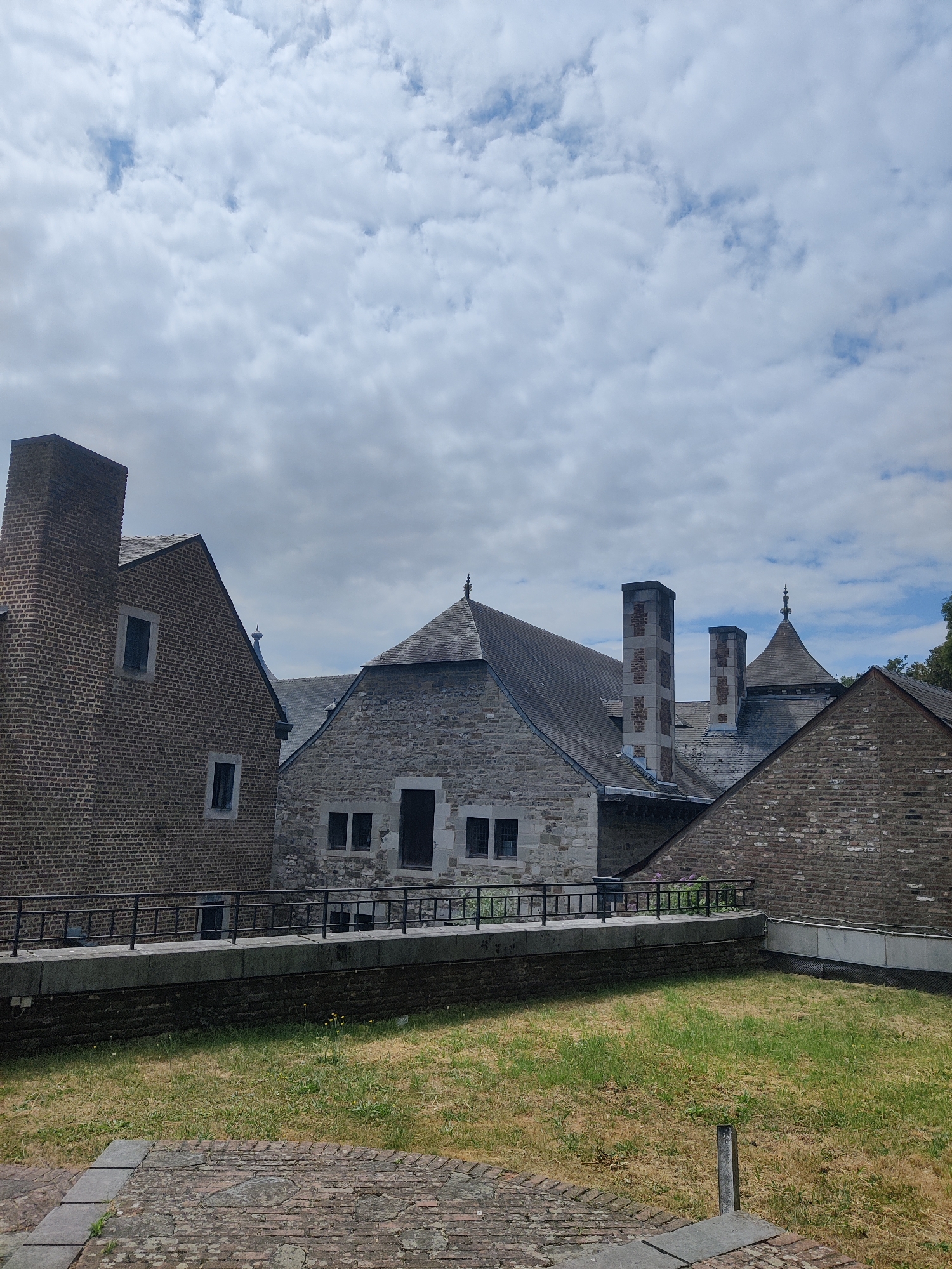 The image shows an exterior view of several historical buildings, likely part of a castle or a similar complex, under a partly cloudy sky. The architecture features brick and stone construction with pitched roofs. Chimneys of various designs are prominent on the rooftops. A black metal railing runs along a walkway in the foreground, separating it from a grassy area below. The ground in the immediate foreground is paved with bricks. The scene appears to be a public space or historical site.
The buildings depicted are part of the **Curtius Museum**, located at the **Quai de Maestricht 13, 4000 Liège, Belgium.** The museum complex is situated on a hill overlooking the Meuse River.