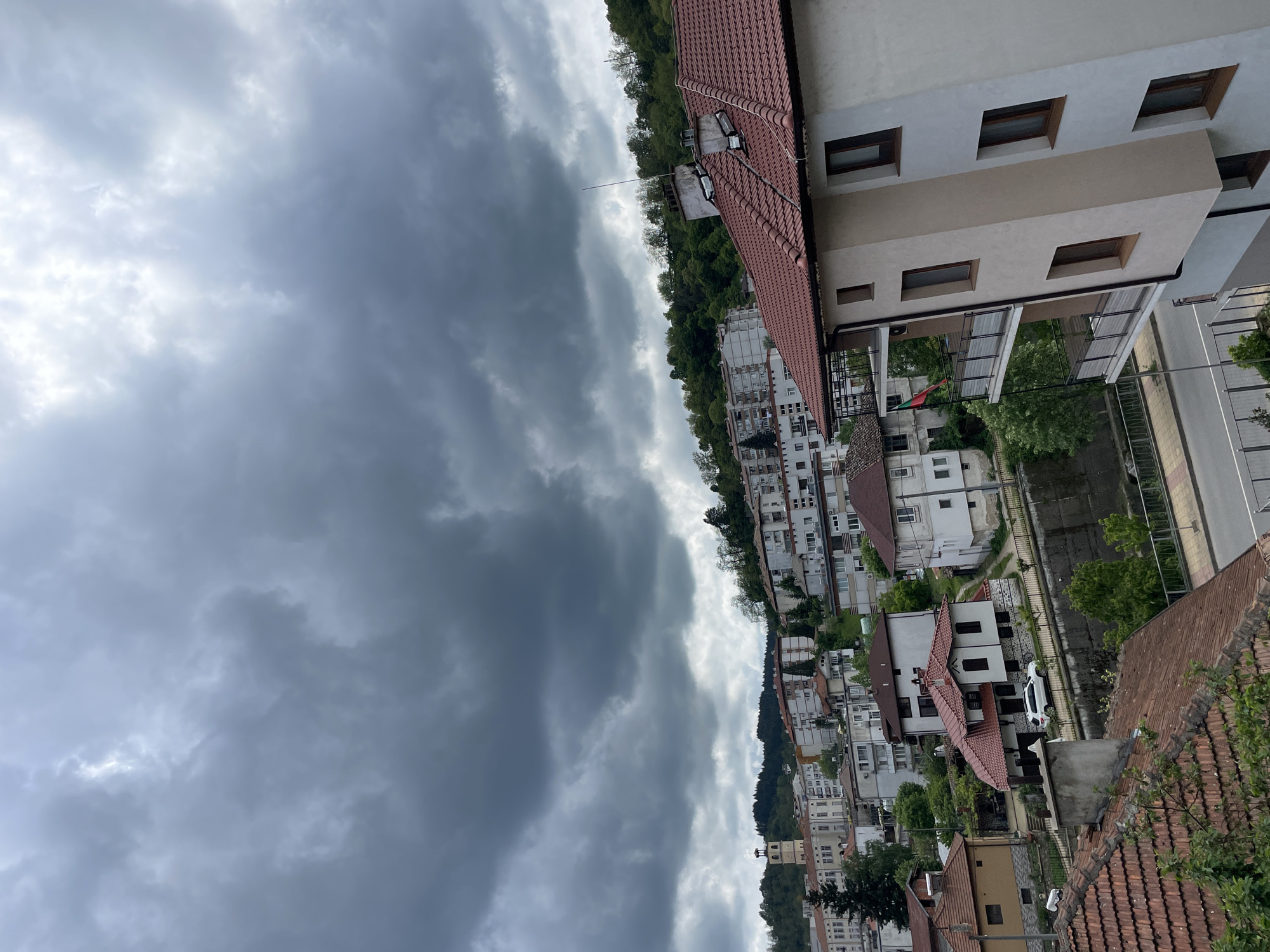 The image depicts a view of Zlatograd, Bulgaria, under a heavily clouded sky. The residential area is composed of multi-story buildings with red-tiled roofs, nestled amongst trees. A white car is visible parked near one of the houses. The sky is dominated by dense, dark clouds. A road is visible to the right.