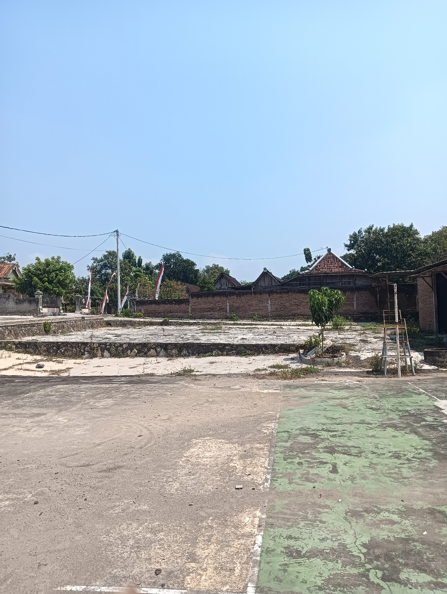 A daytime, eye-level shot captures an outdoor scene in Tanggan, Indonesia. The foreground is dominated by an aged, cracked asphalt surface, part of which is painted green. Beyond this, a raised, concrete area with rough stone edging is visible, covered in sparse vegetation and debris. In the background, a brick wall runs horizontally, behind which traditional Indonesian houses with red-tiled roofs are nestled amongst lush green trees. Several Indonesian flags, red and white, are displayed on poles, suggesting a local celebration or commemoration. A simple metal ladder stands near a covered structure on the right. The sky above is a clear, pale blue.
