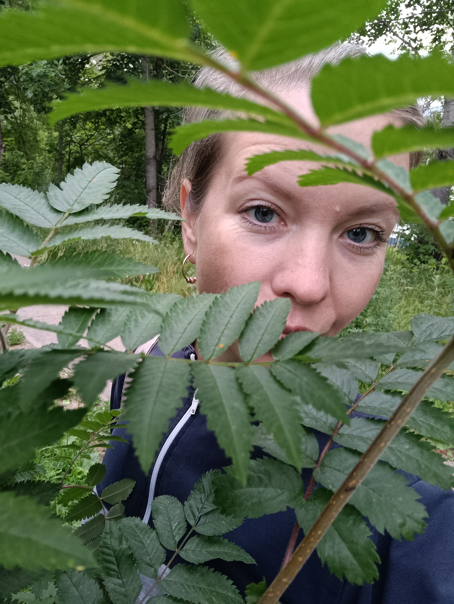 A woman is peering through foliage in a park in Chaikovsky, Russia. She has fair skin, blue eyes, and light-colored hair. She is wearing a dark blue zippered jacket. The background consists of green trees and grass.